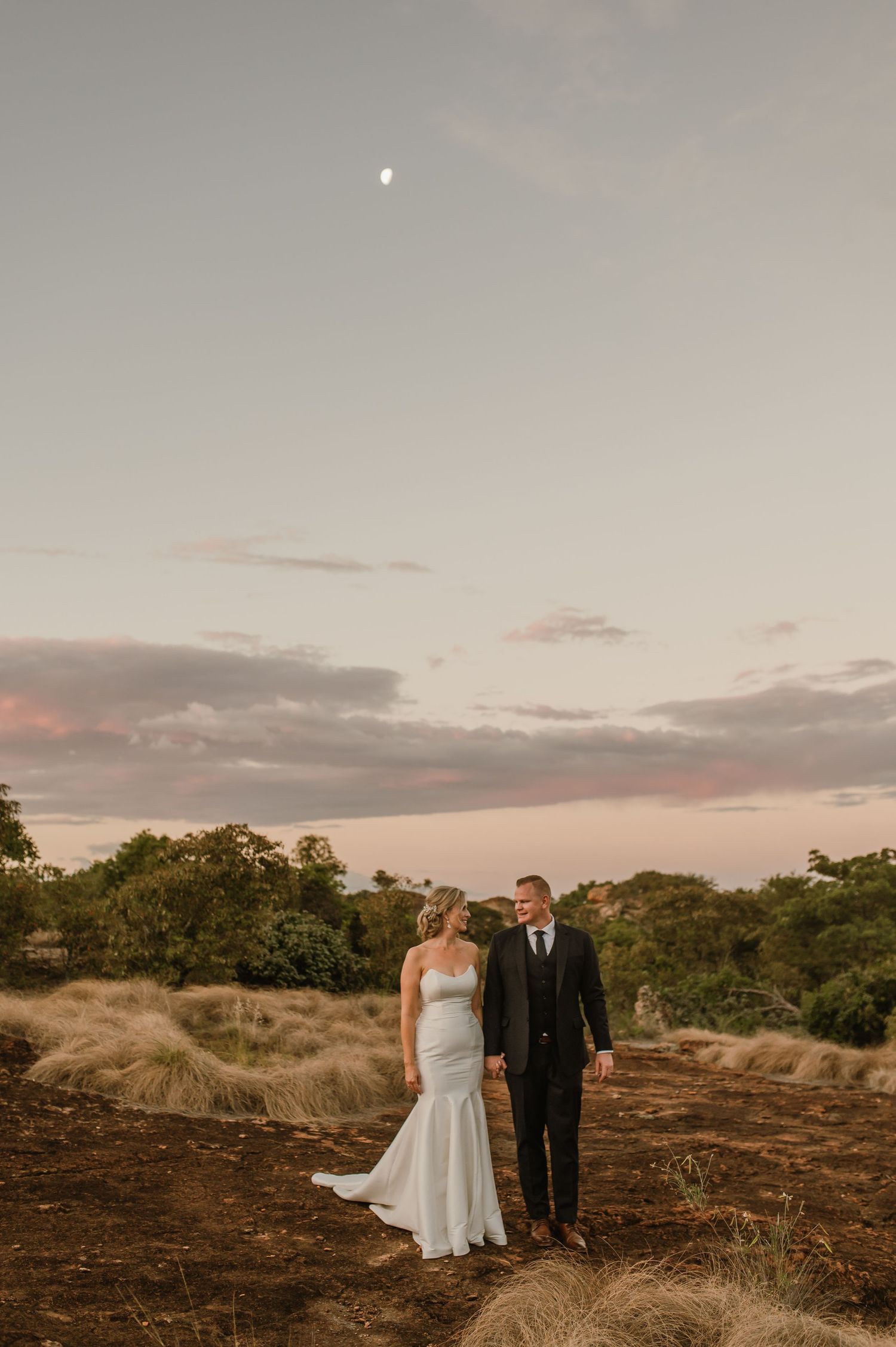 Newlyweds stand together against a scenic sunset backdrop on rocky terrain.