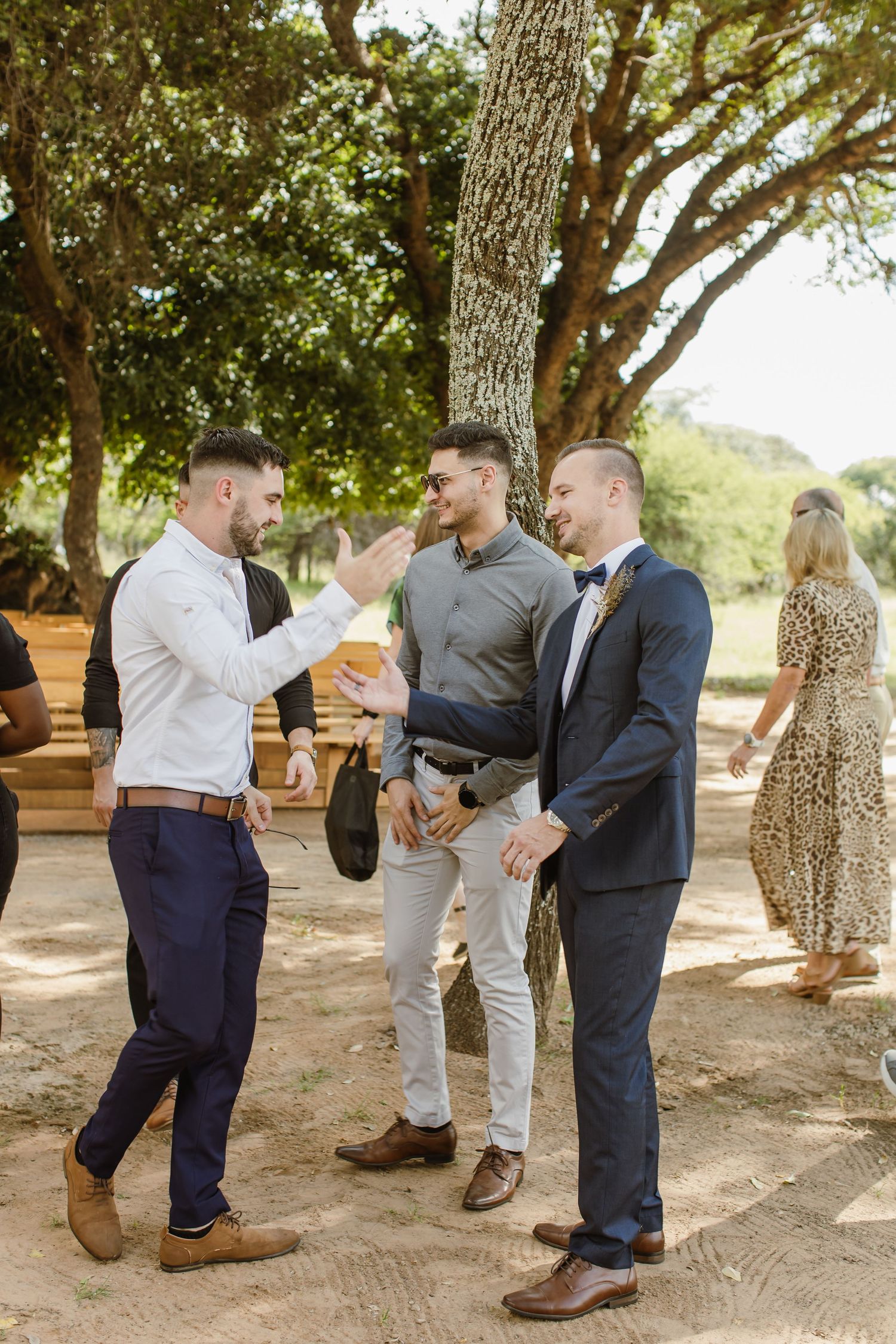 Groomsmen sharing celebratory moments under oak trees at an outdoor venue.