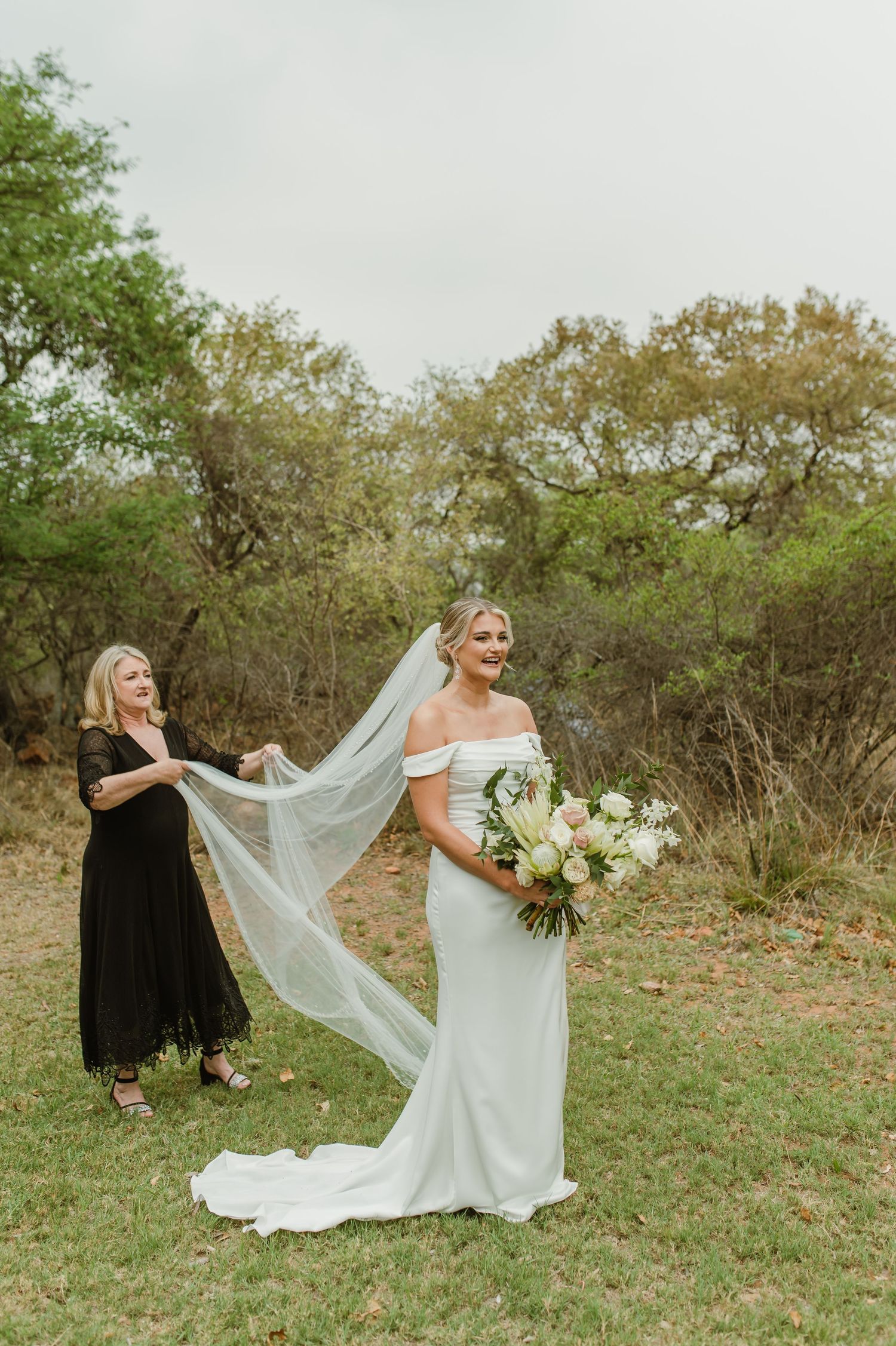 Elegant bride in white dress poses with flowing veil in outdoor garden setting while bridesmaid assists.