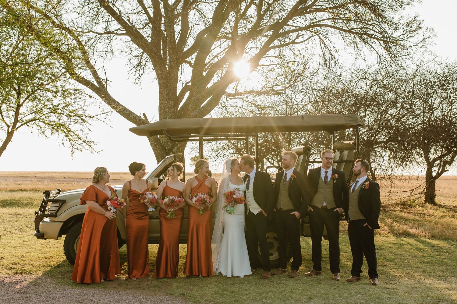 Wedding party poses in a line under trees at sunset with bridesmaids in rust-colored dresses.