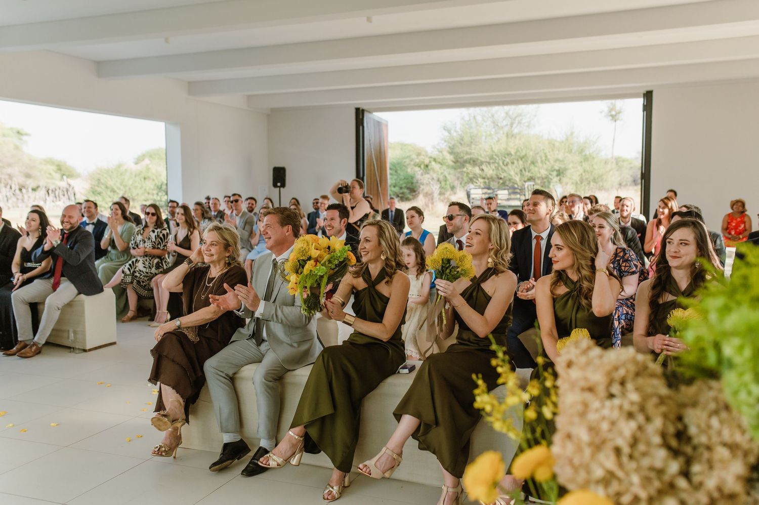 Wedding guests seated in rows at an indoor ceremony venue with natural lighting and floral decorations.