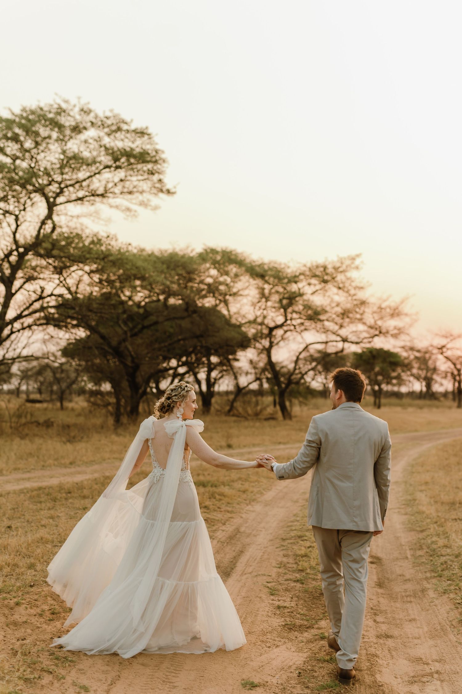 Newlyweds walking hand in hand down dirt path at sunset in African savanna.