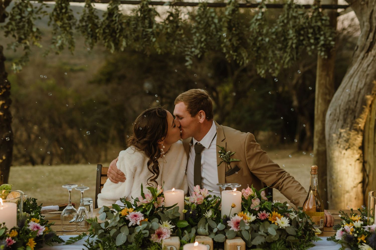 Romantic outdoor wedding reception under trees with string lights and floral decorations.