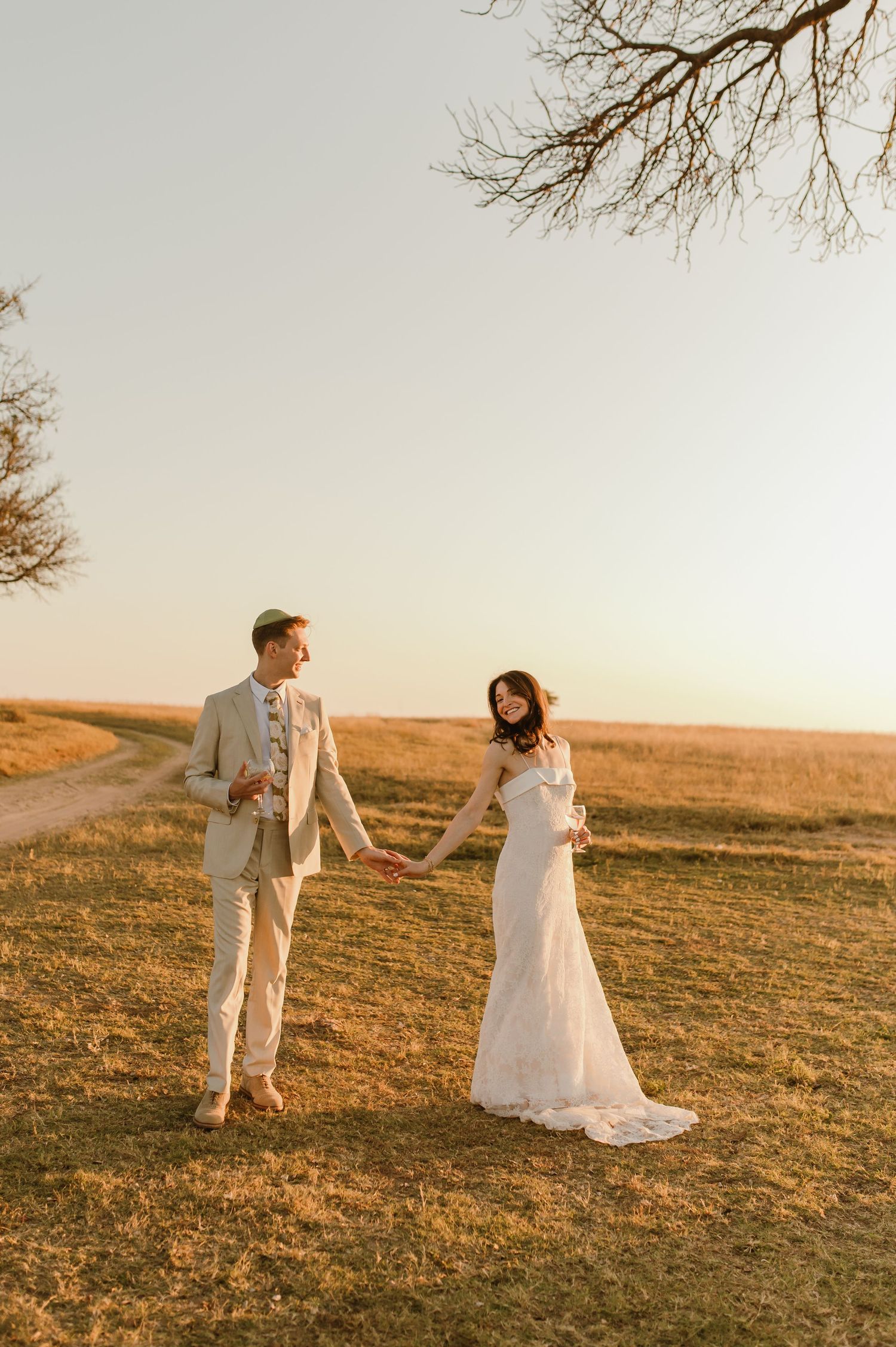 Bride in white lace gown and groom in tan suit hold hands in golden field at sunset.
