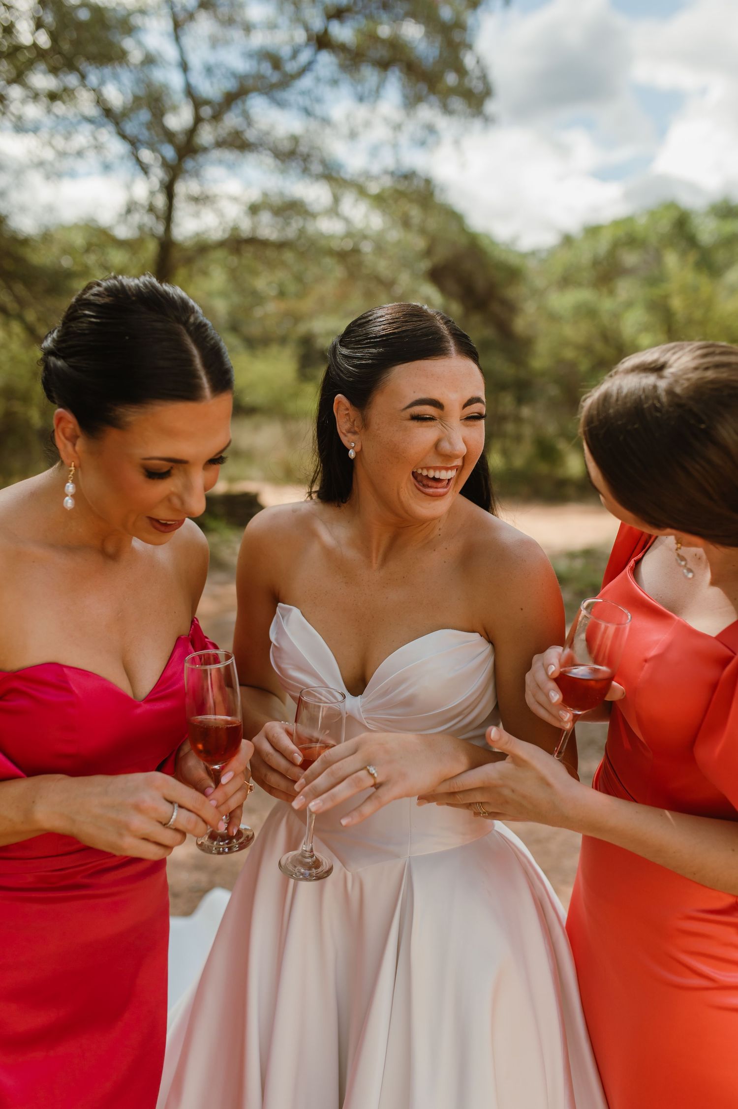 Bride in white strapless gown laughing with two bridesmaids in red dresses holding champagne.