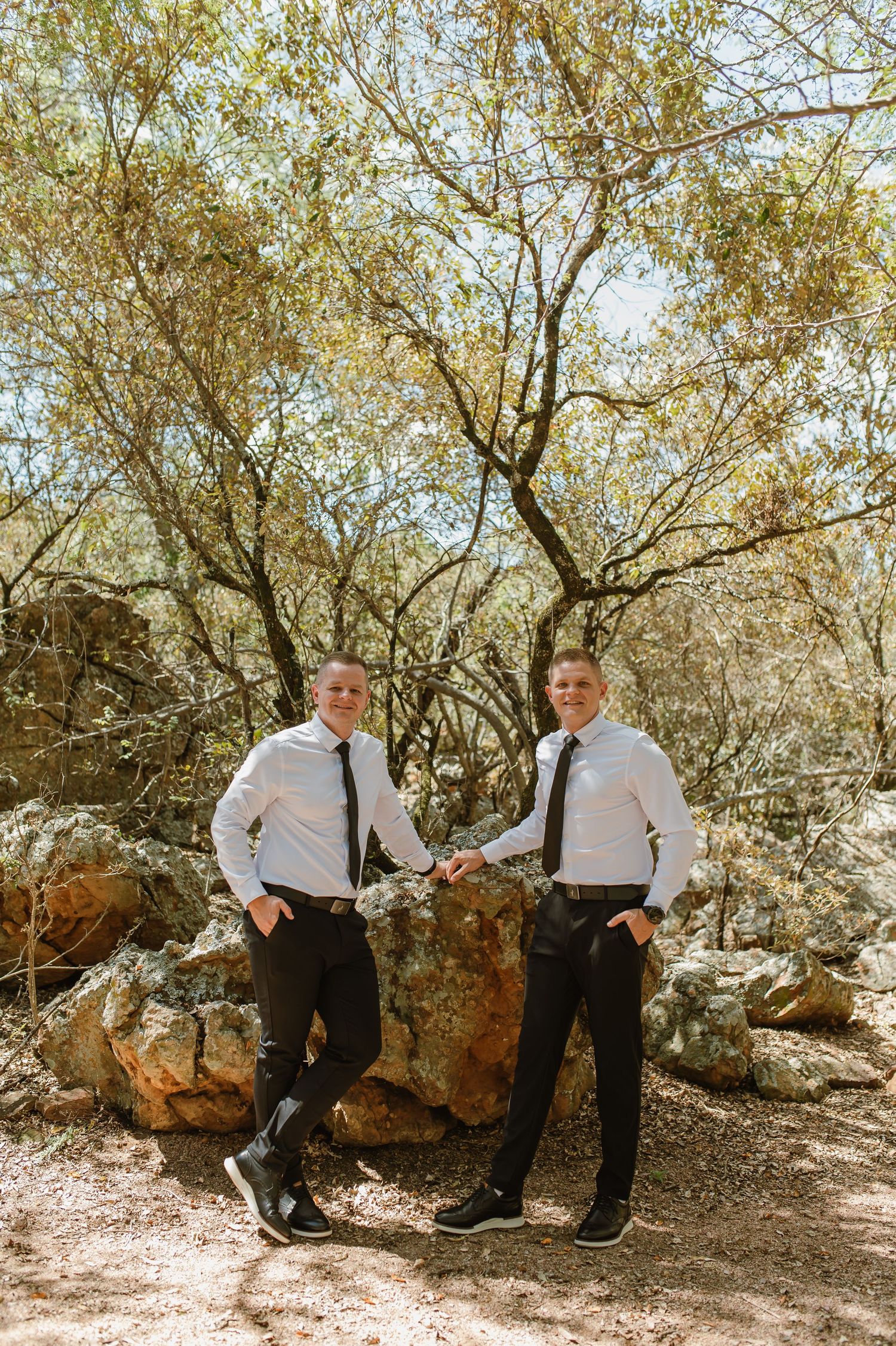 Two groomsmen in white shirts and black ties posing confidently against large rocks in a natural woodland setting.