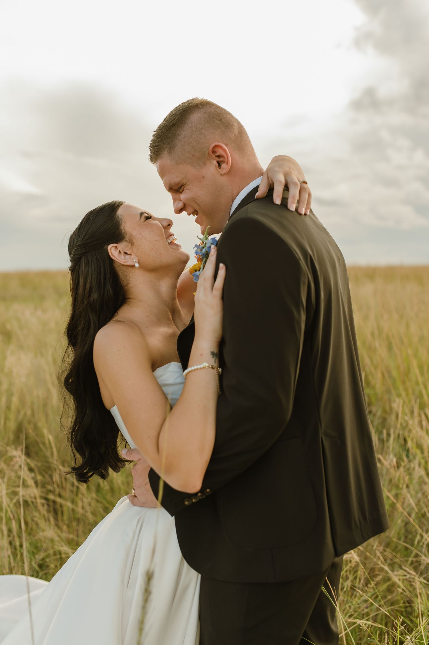 Bride and groom sharing a romantic embrace in a golden grass field under a cloudy sky after their wedding.