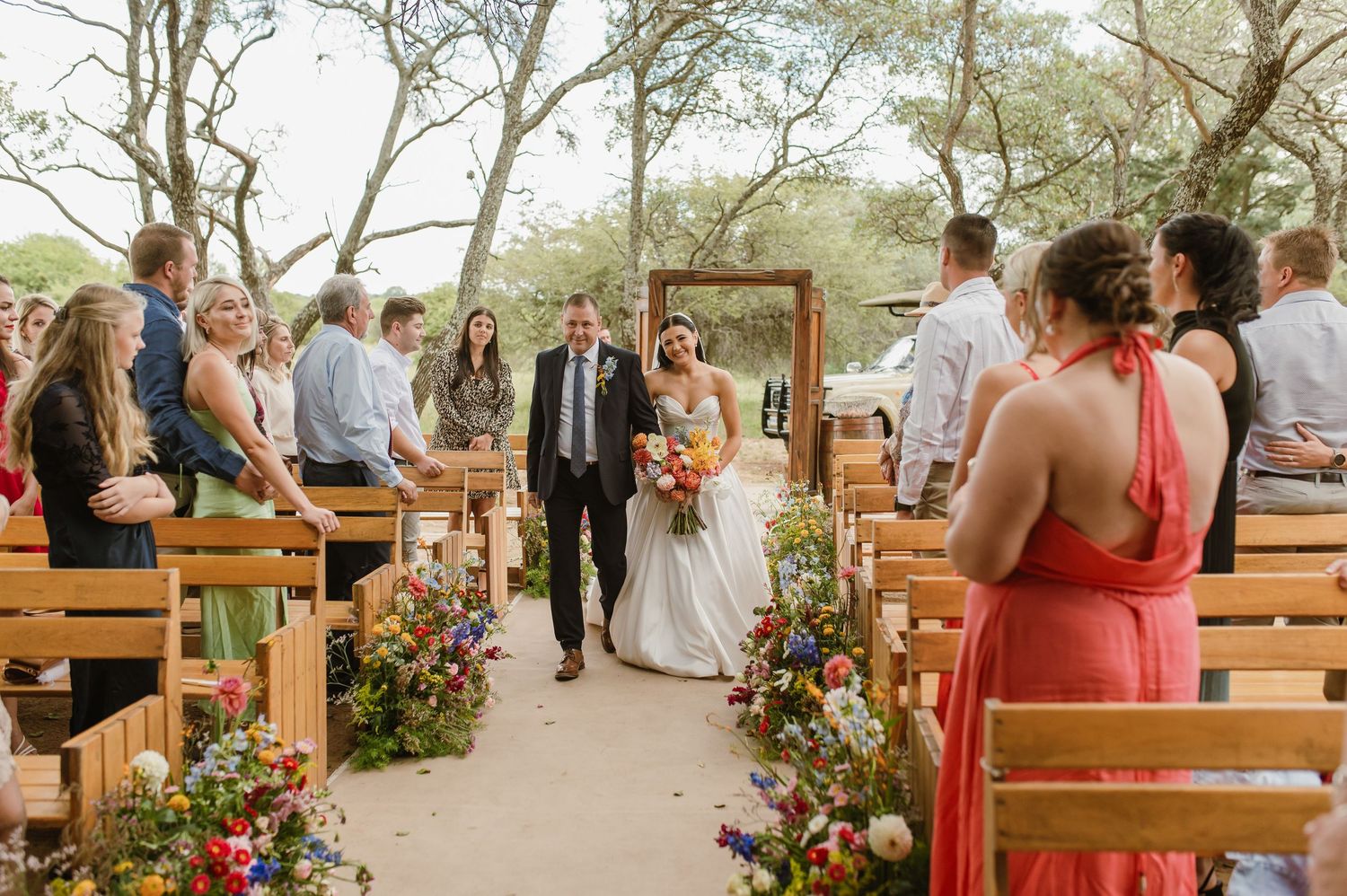 Bride walking down the aisle with her father at an outdoor wedding ceremony lined with colorful wildflowers.