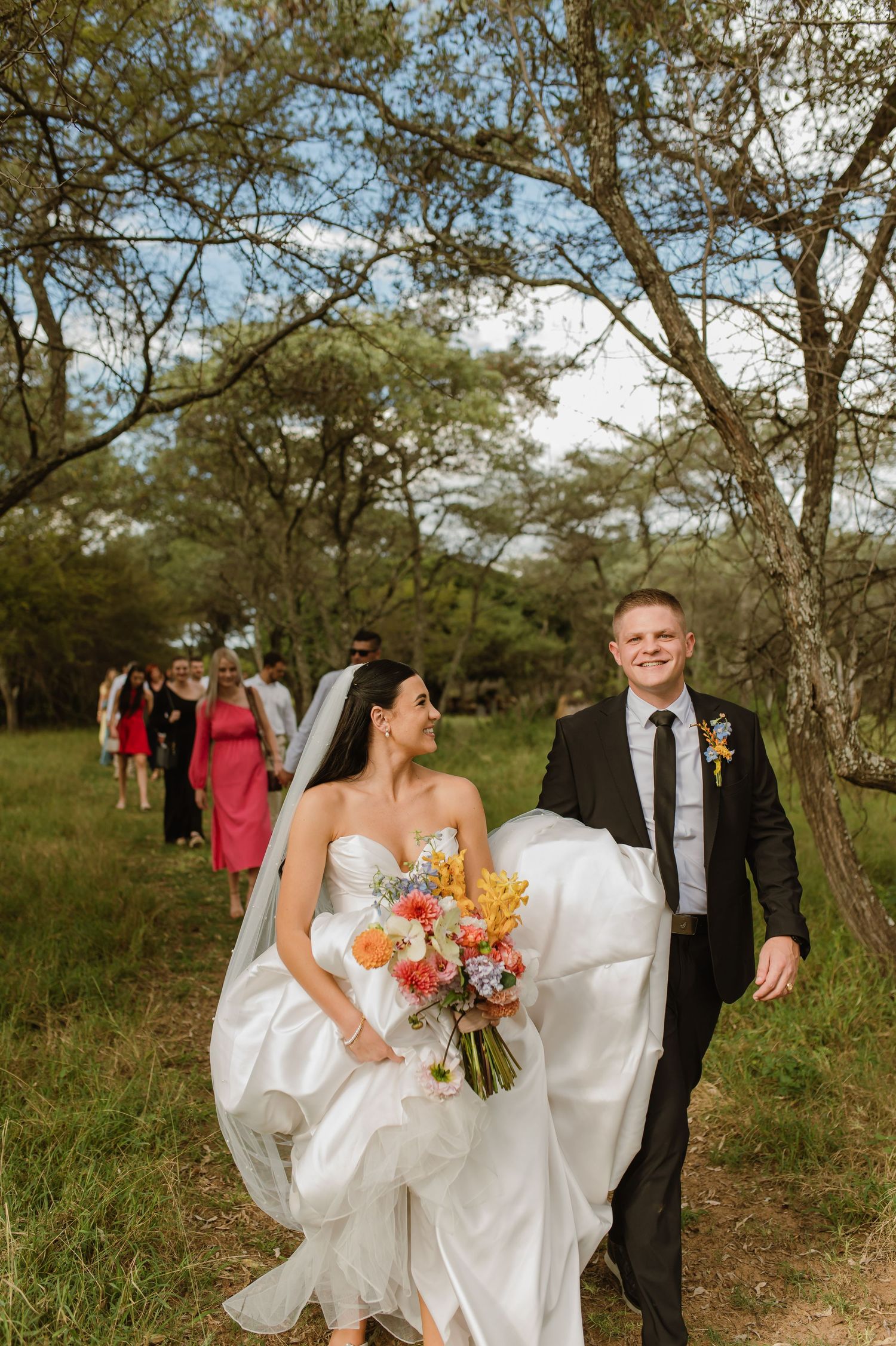 Joyful newlyweds walking through trees with wedding guests following after outdoor bush ceremony.