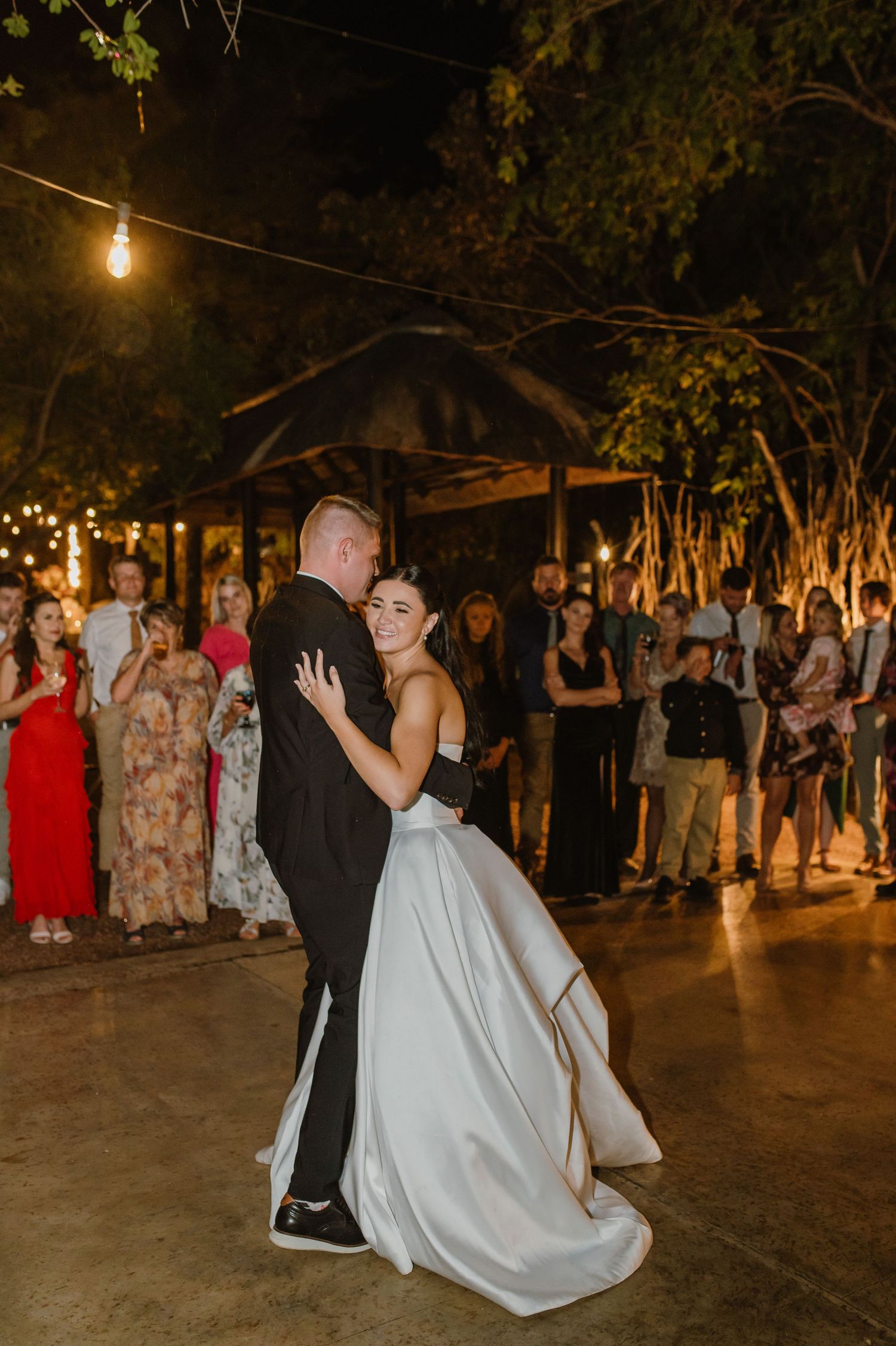 Bride and groom share first dance outdoors at night under string lights with guests watching.