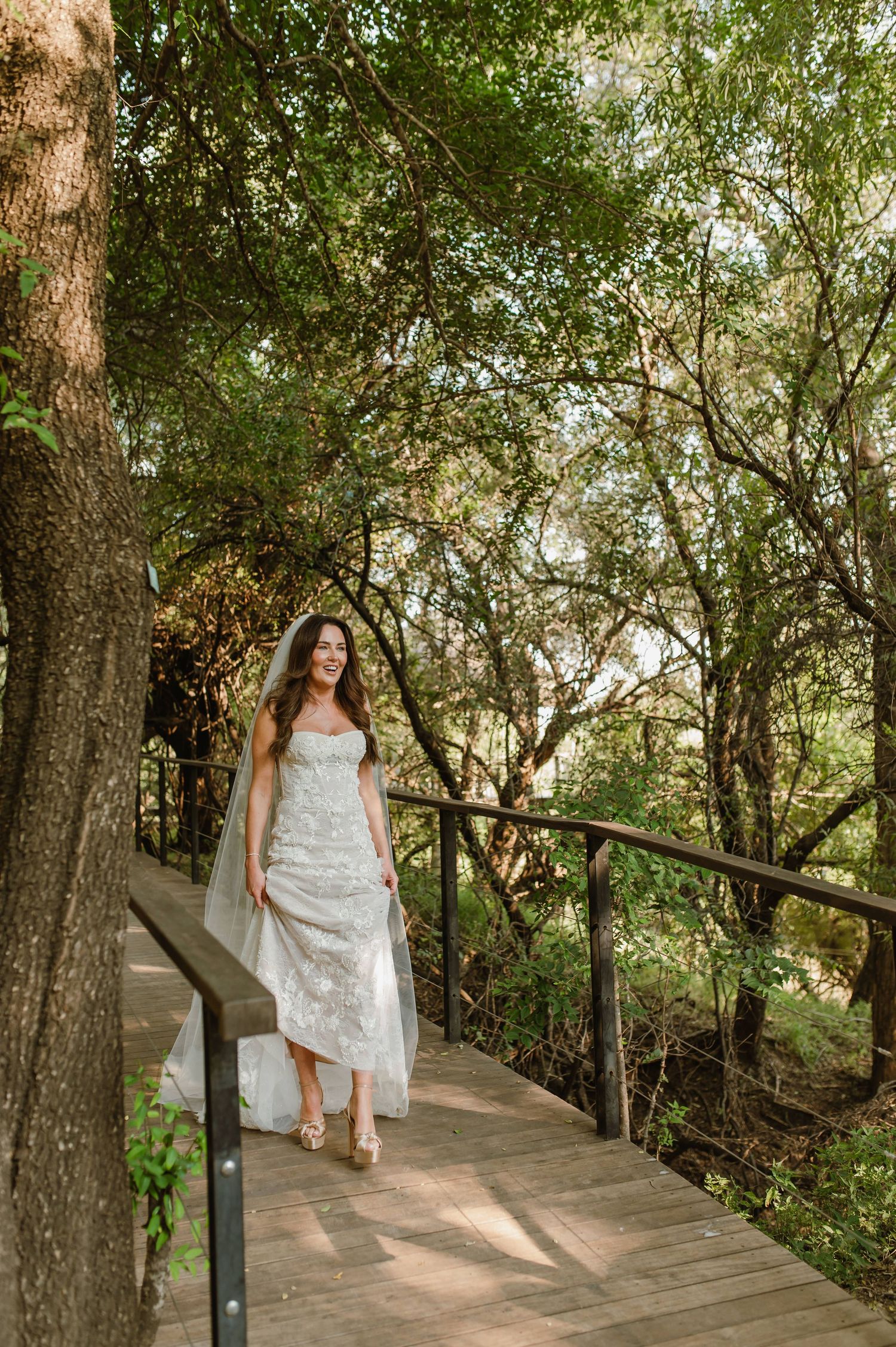 Radiant bride in lace wedding gown and veil walking on wooden bridge surrounded by lush trees.