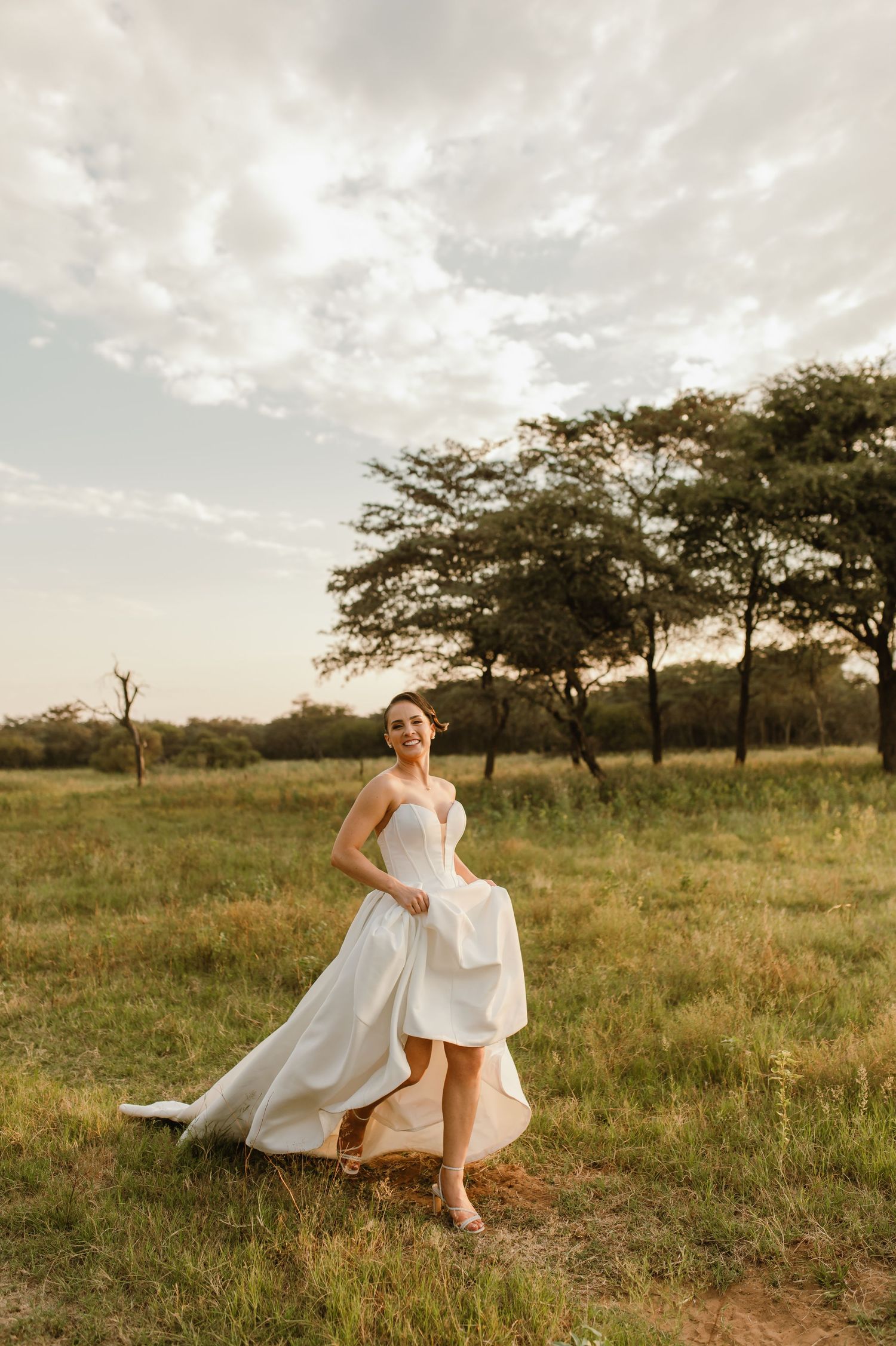 A bride in a flowing white dress twirls in a sunlit savanna field during golden hour.