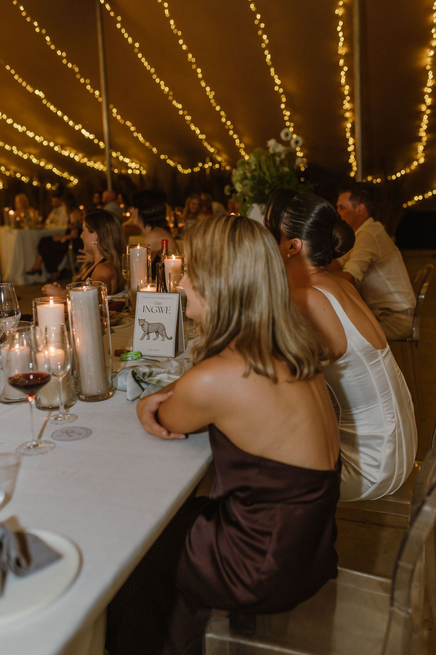 Wedding reception dinner under twinkling string lights with guests seated at long tables with white linens and candles.