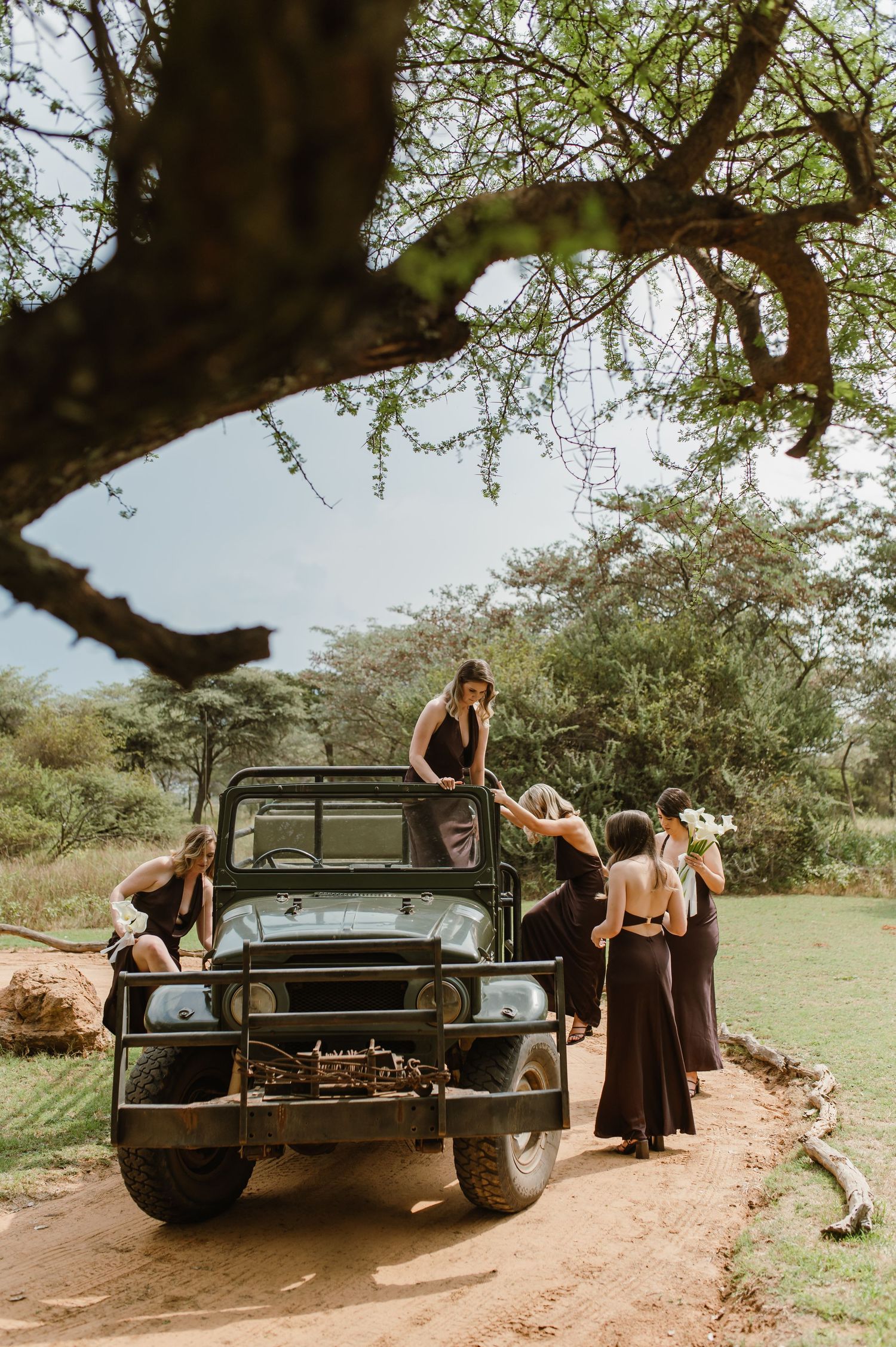 Wedding party gathered around vintage black off-road vehicle in rustic outdoor setting.