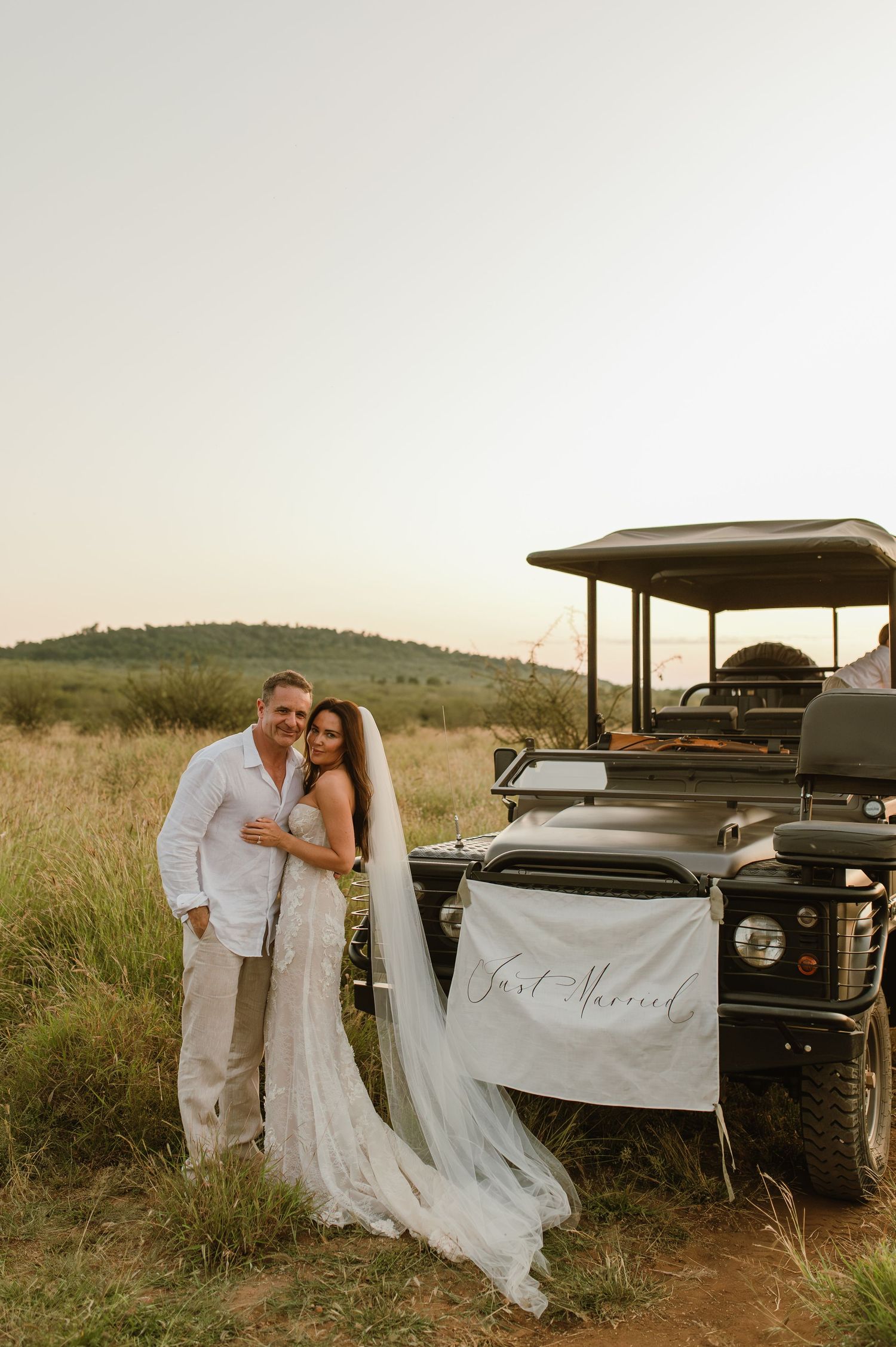 Newlyweds pose beside Just Married safari vehicle in open savanna, bride in lace gown with long veil.