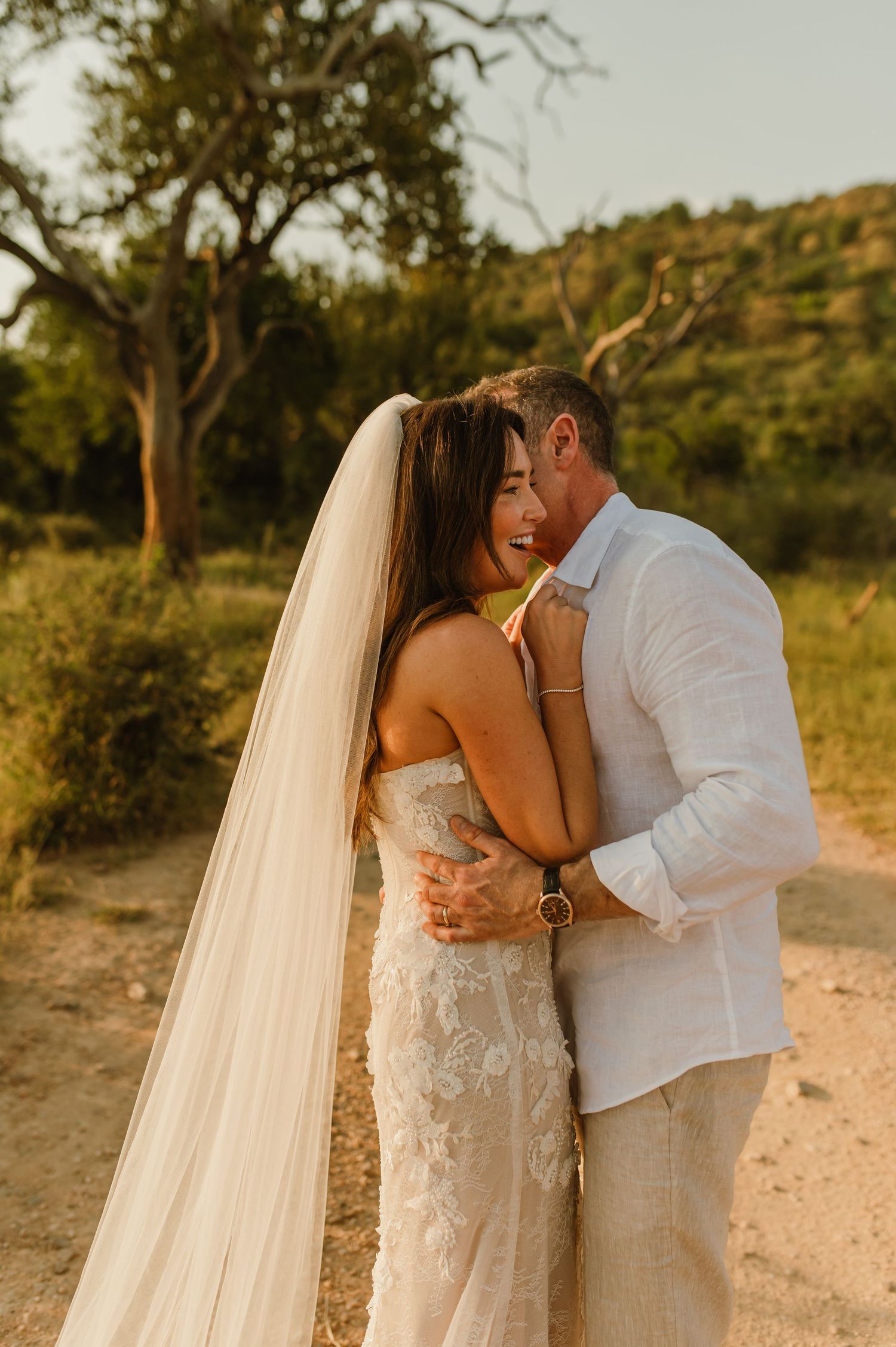 Bride and groom embrace at golden hour, she laughs in lace strapless gown with veil, he wears white linen shirt.