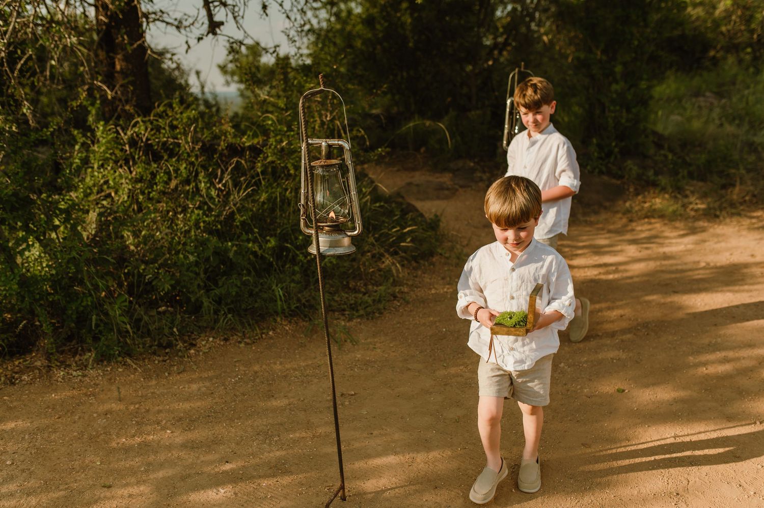 Two young boys in white outfits walk along a sandy path surrounded by lush greenery near a vintage lantern.