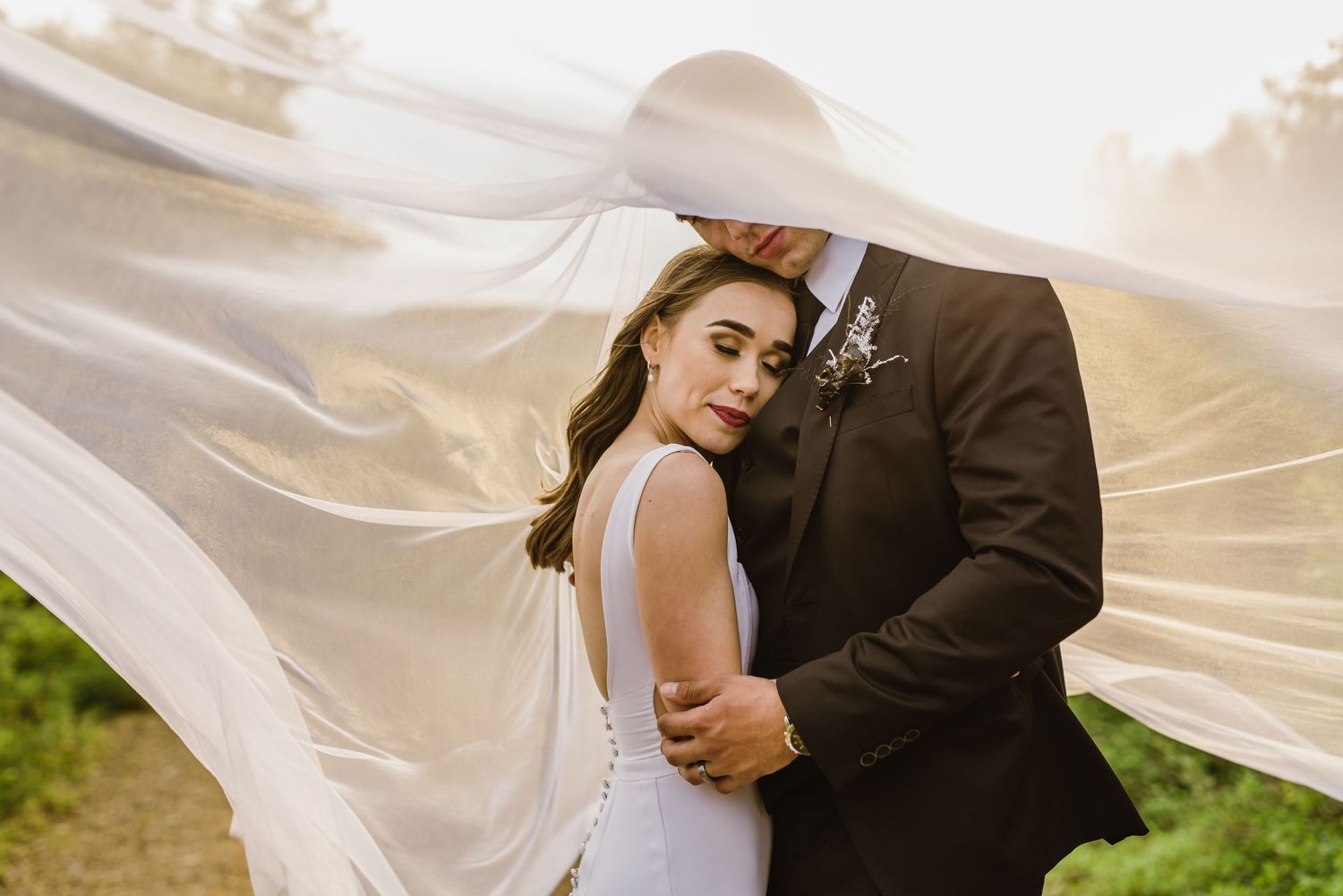 Romantic wedding couple embraces with flowing veil against sunset sky in outdoor setting.