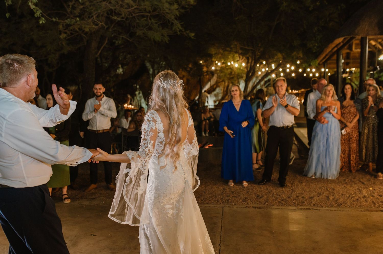 Couple dancing on outdoor patio surrounded by guests and string lights at evening celebration.