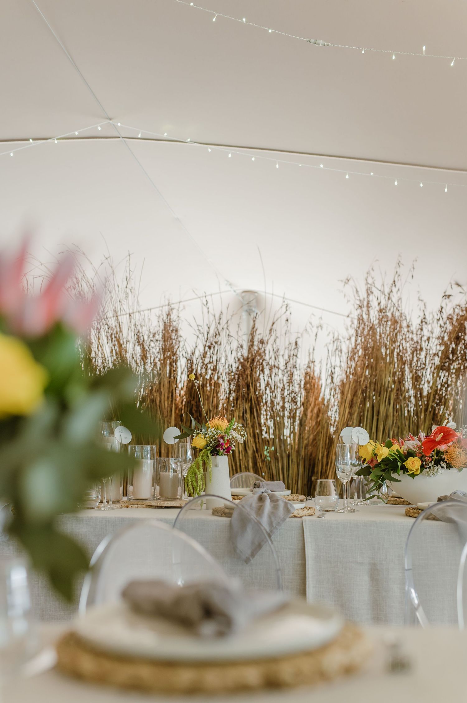 Elegant wedding reception table settings with woven chargers, white linens, and dried grass decor in the background.