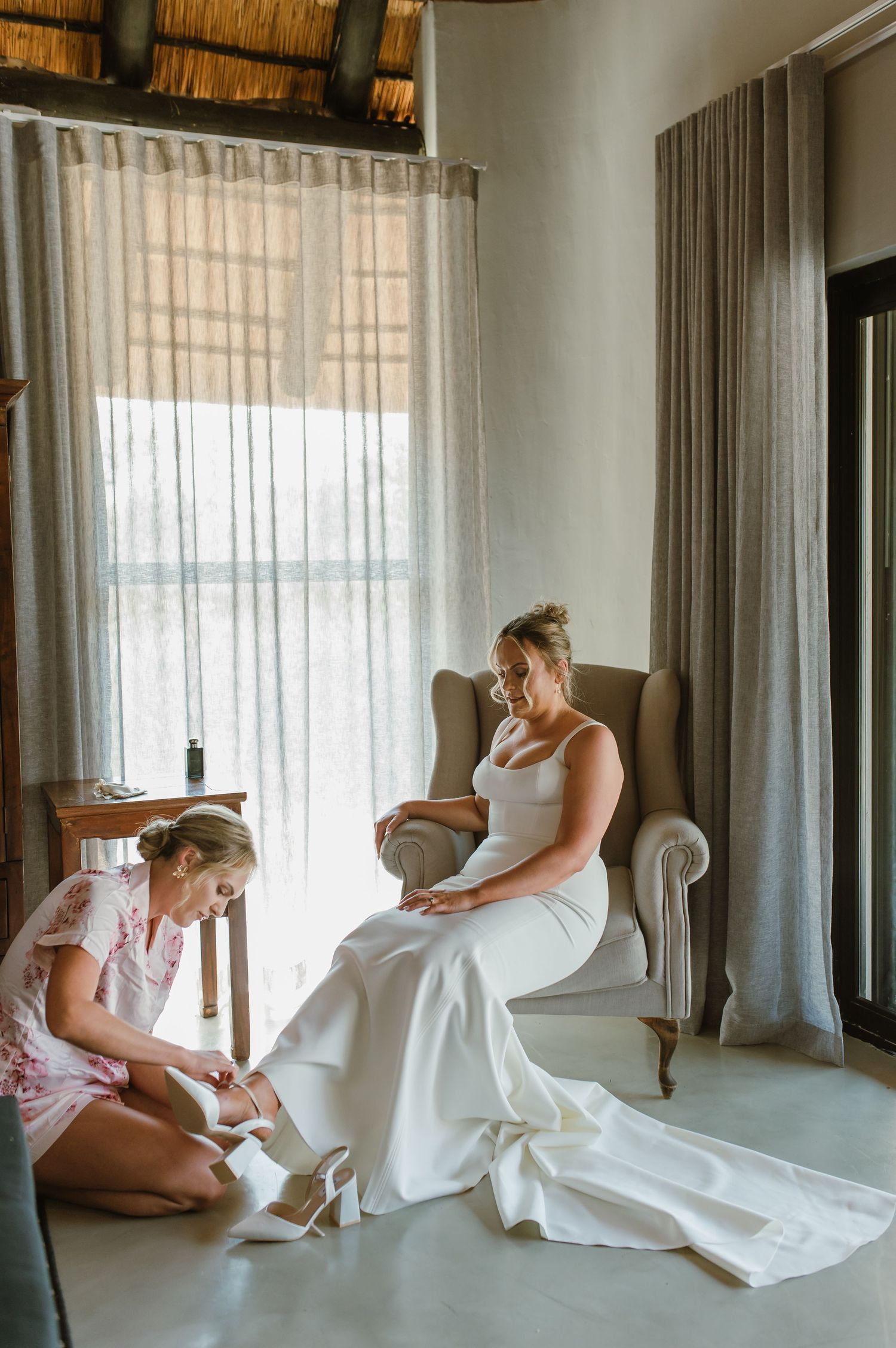 A person in a white wedding dress kneels on an upholstered chair in an elegant room.