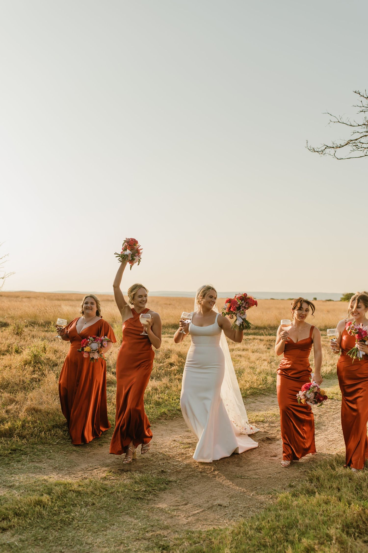 Bridesmaids in terracotta dresses dance and celebrate in a golden field.