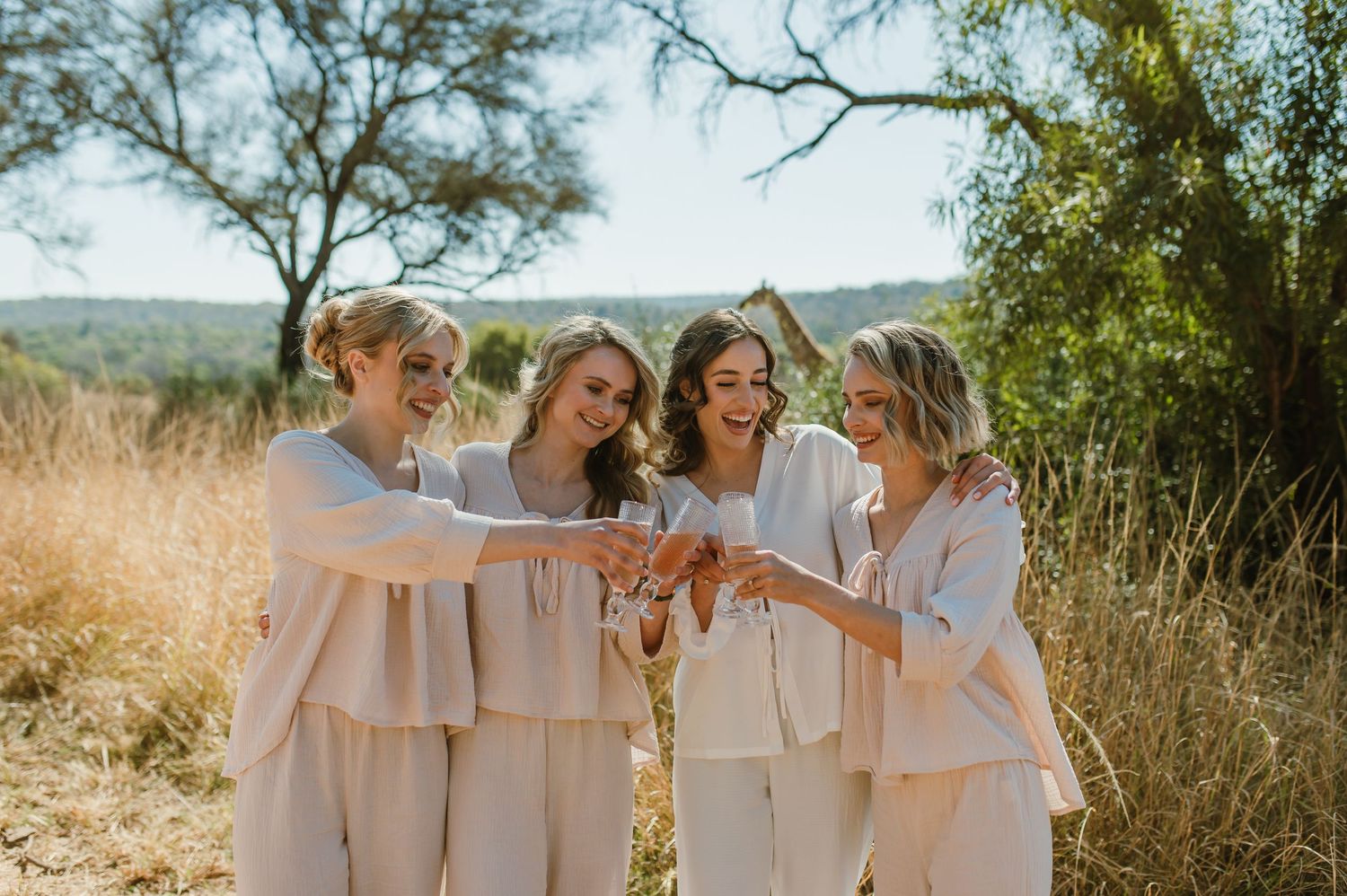A group wearing matching cream-colored dresses laughs together in a sunny outdoor setting with mountains in the background.