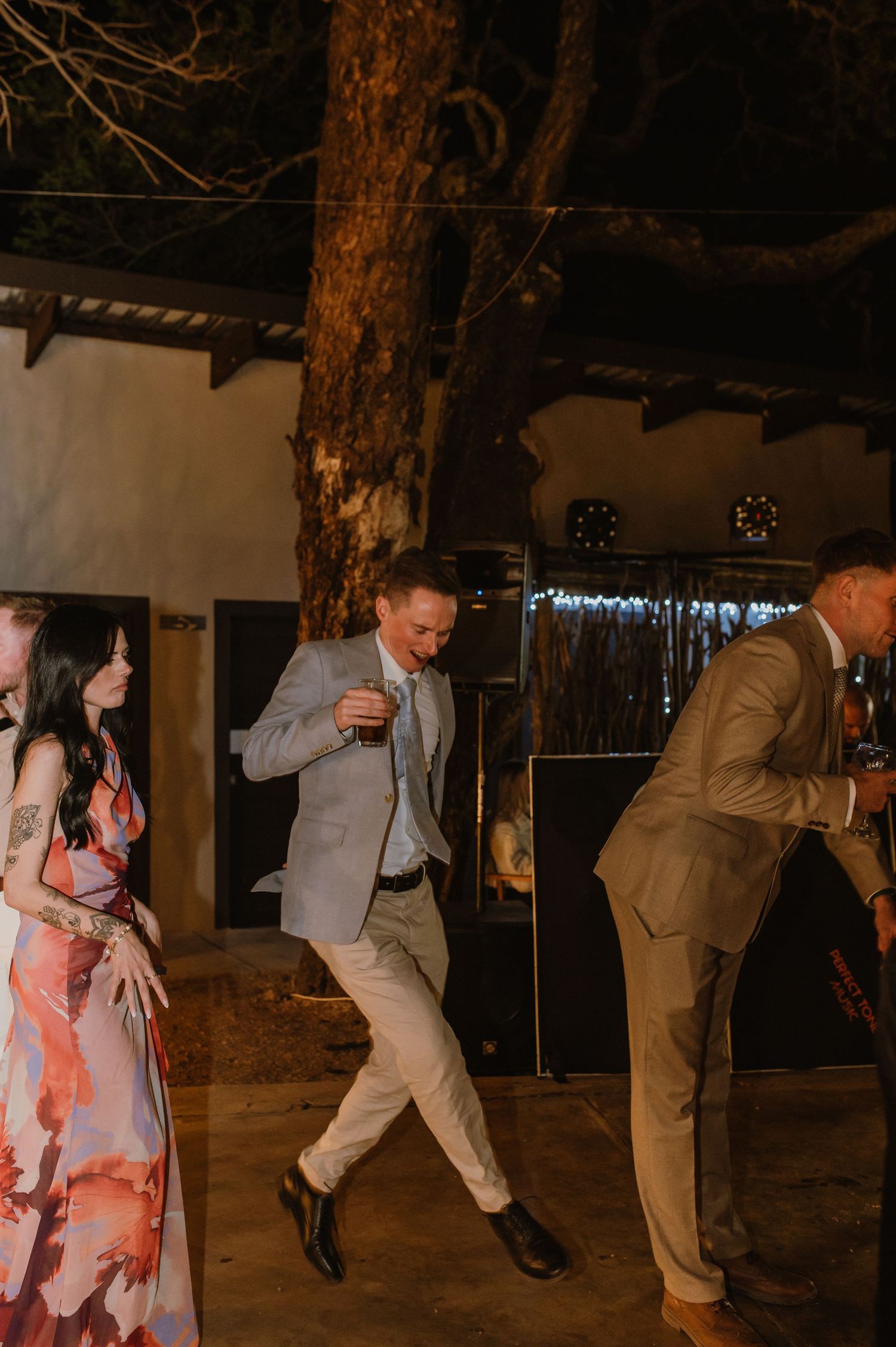 Wedding guests dance and celebrate during a lively reception with people in formal attire and floral dresses.