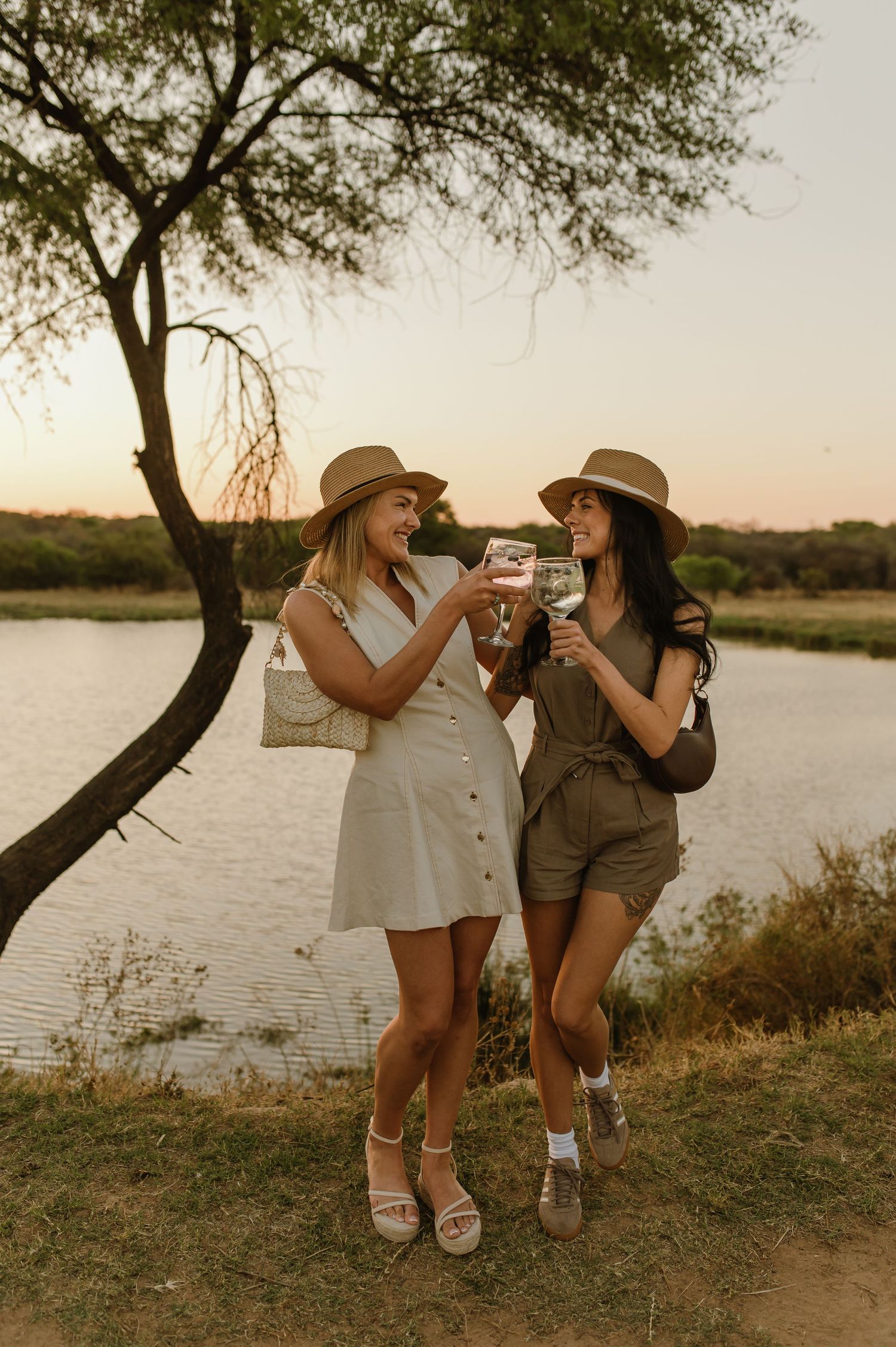 Two friends toasting drinks by the water at sunset wearing safari-inspired outfits.
