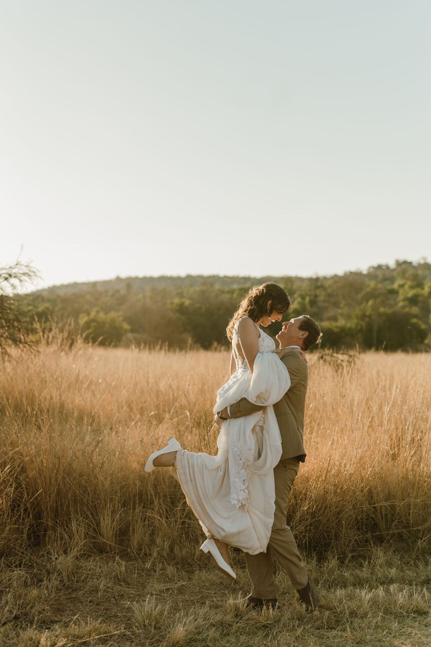 Groom lifts bride in white dress during sunset photoshoot in tall grass field.