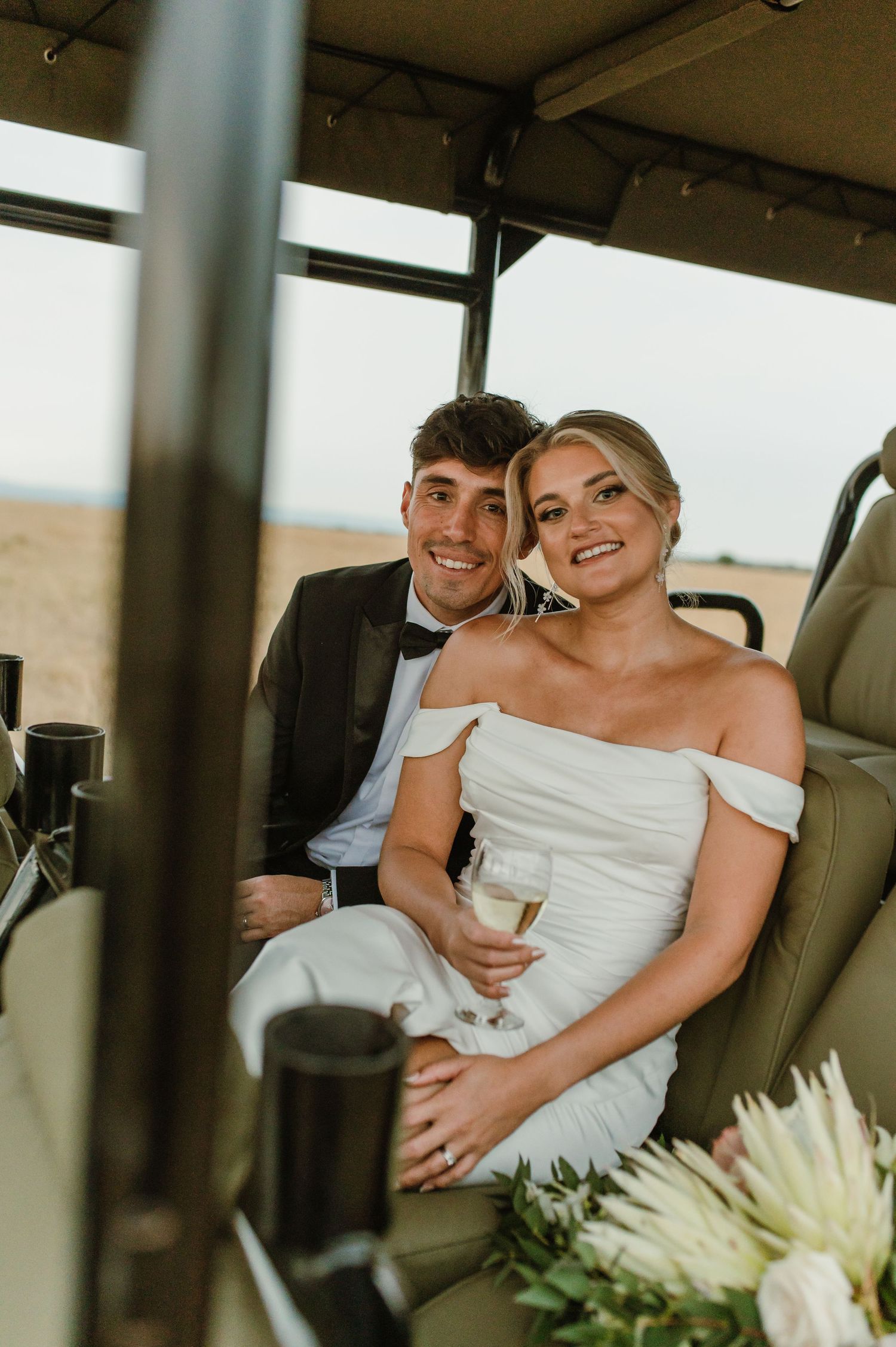 Newlyweds laugh together while seated in a safari-style vehicle with white flowers.