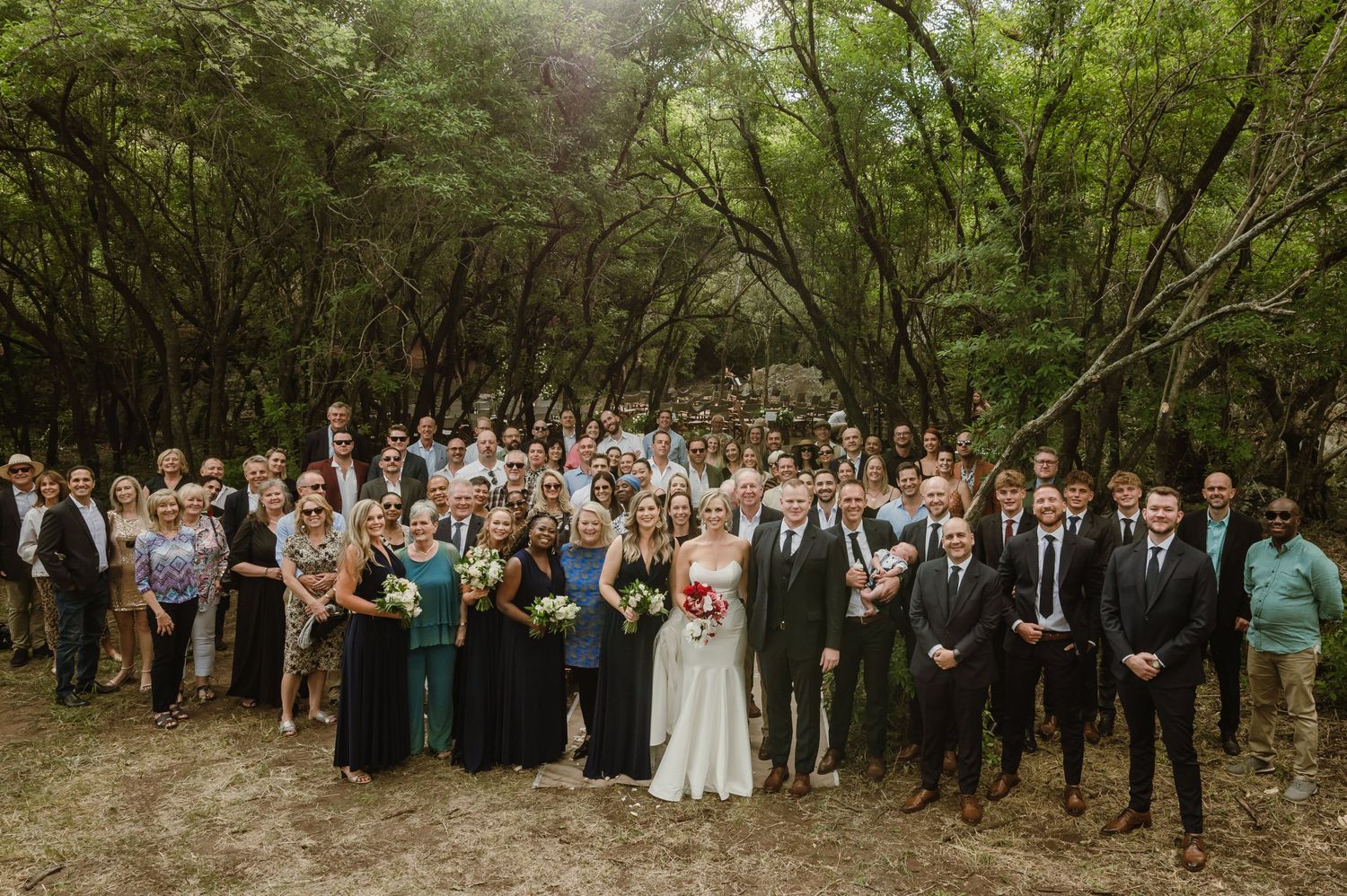 Large group wedding photo of guests and wedding party gathered on a wooded trail.
