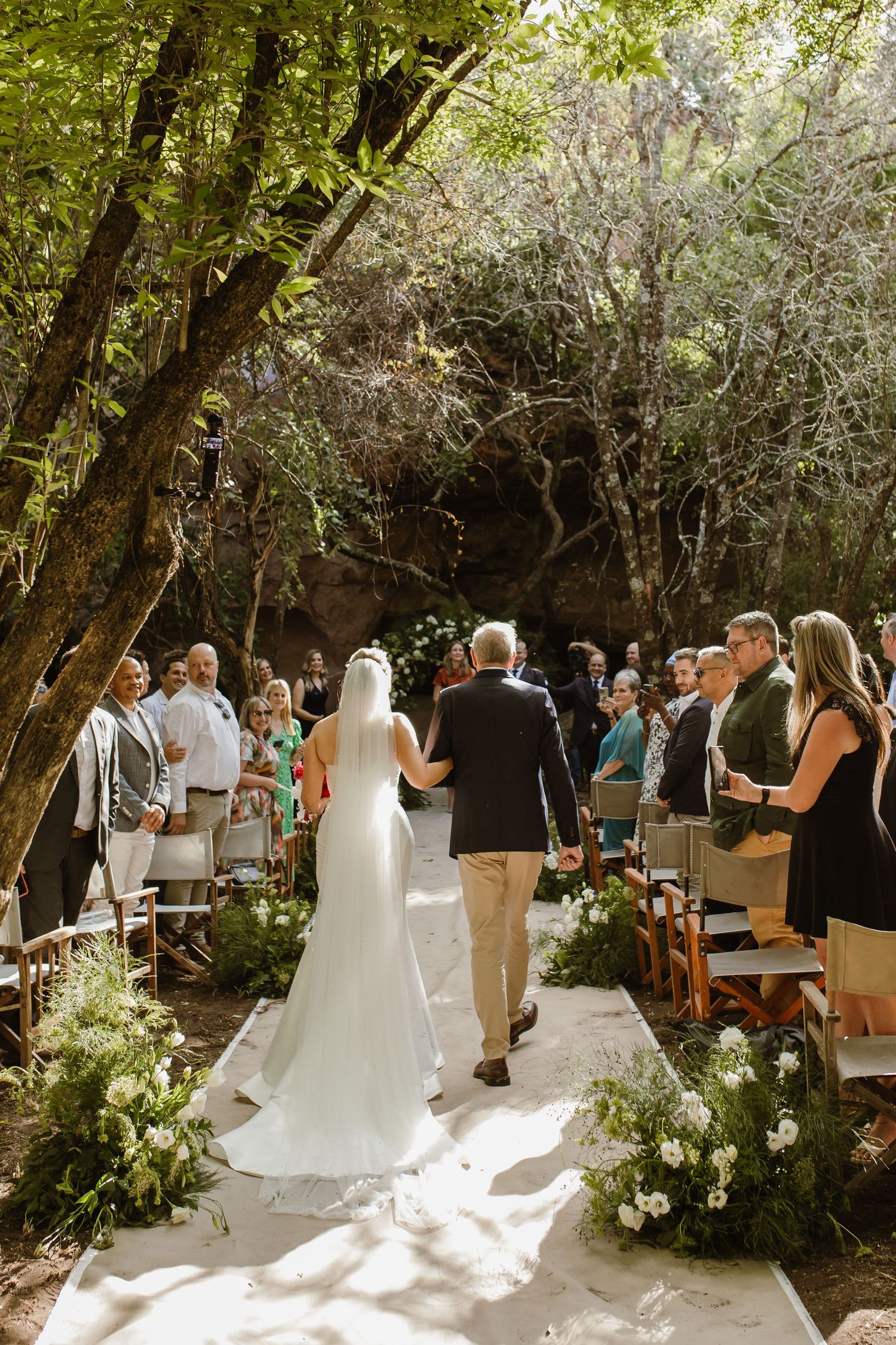 Outdoor wedding ceremony in a wooded setting with white floral decorations and wooden chairs.