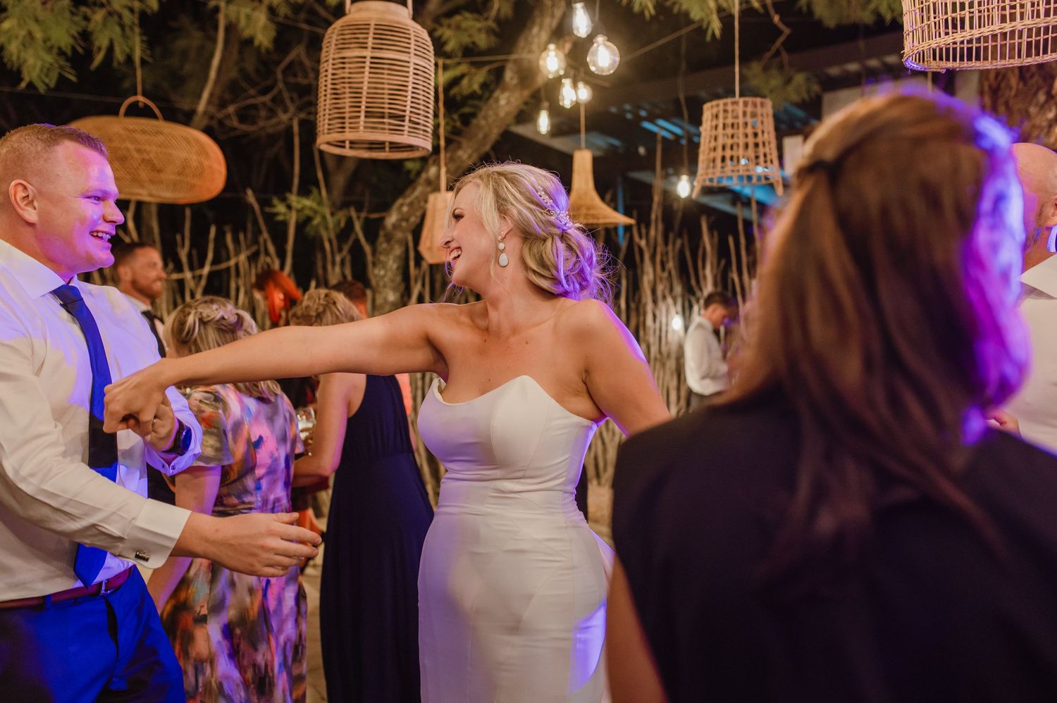 Guests celebrate at nighttime wedding reception under glowing wicker lanterns and purple lighting.