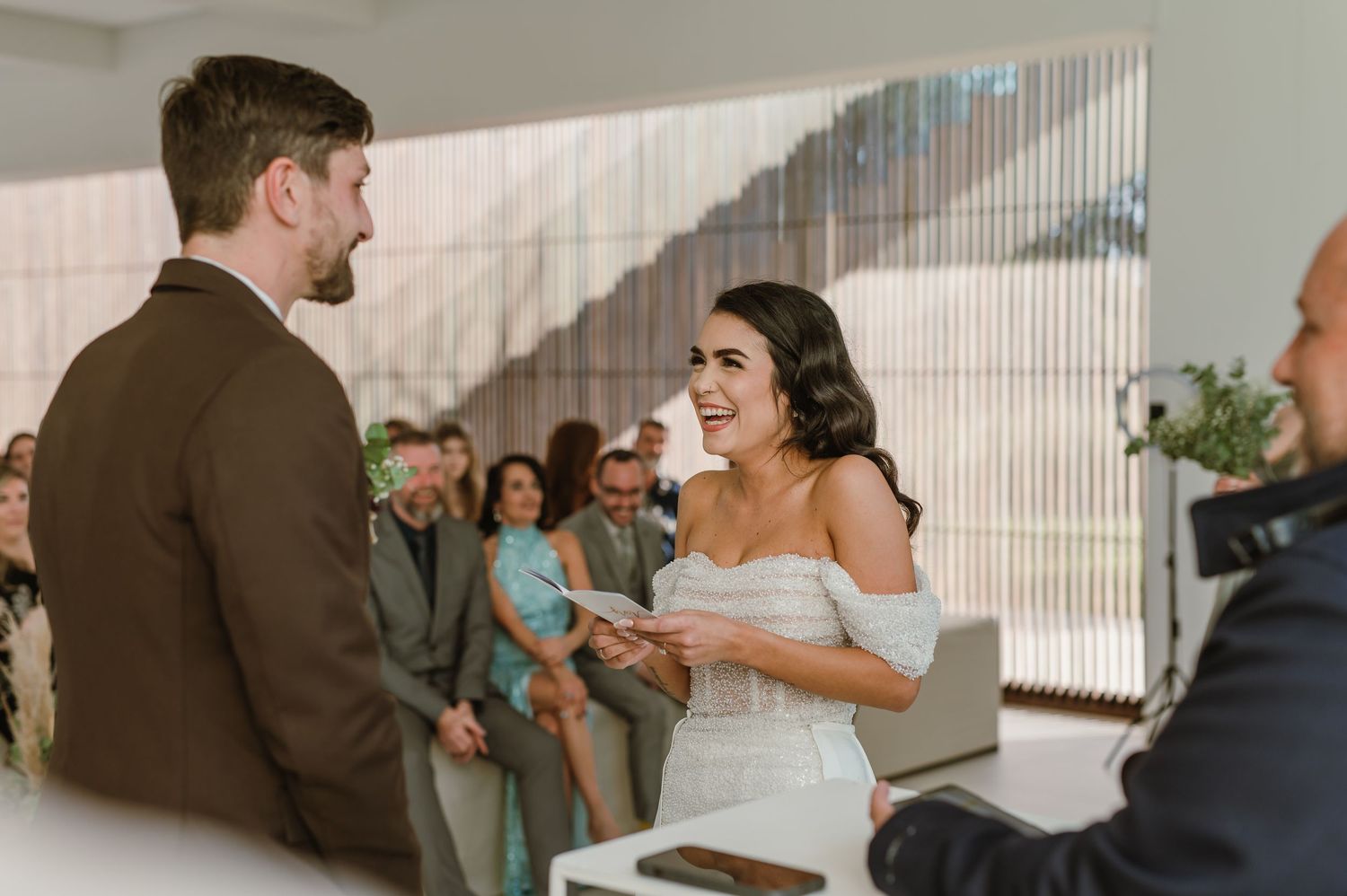 Wedding party members gather in modern white venue with vertical window blinds.