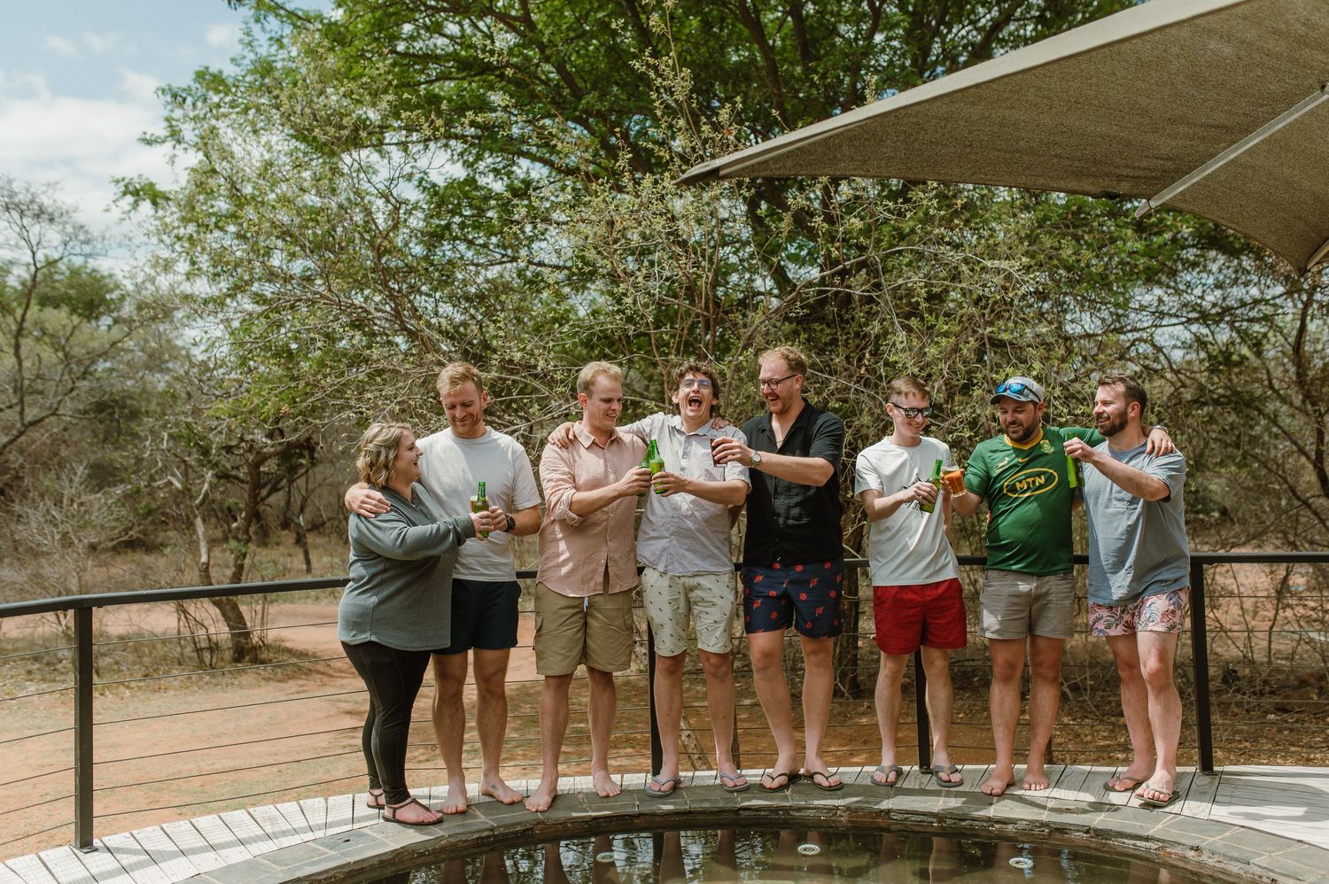 A group of people gathered on a wooden deck overlooking a natural pool in an outdoor setting.