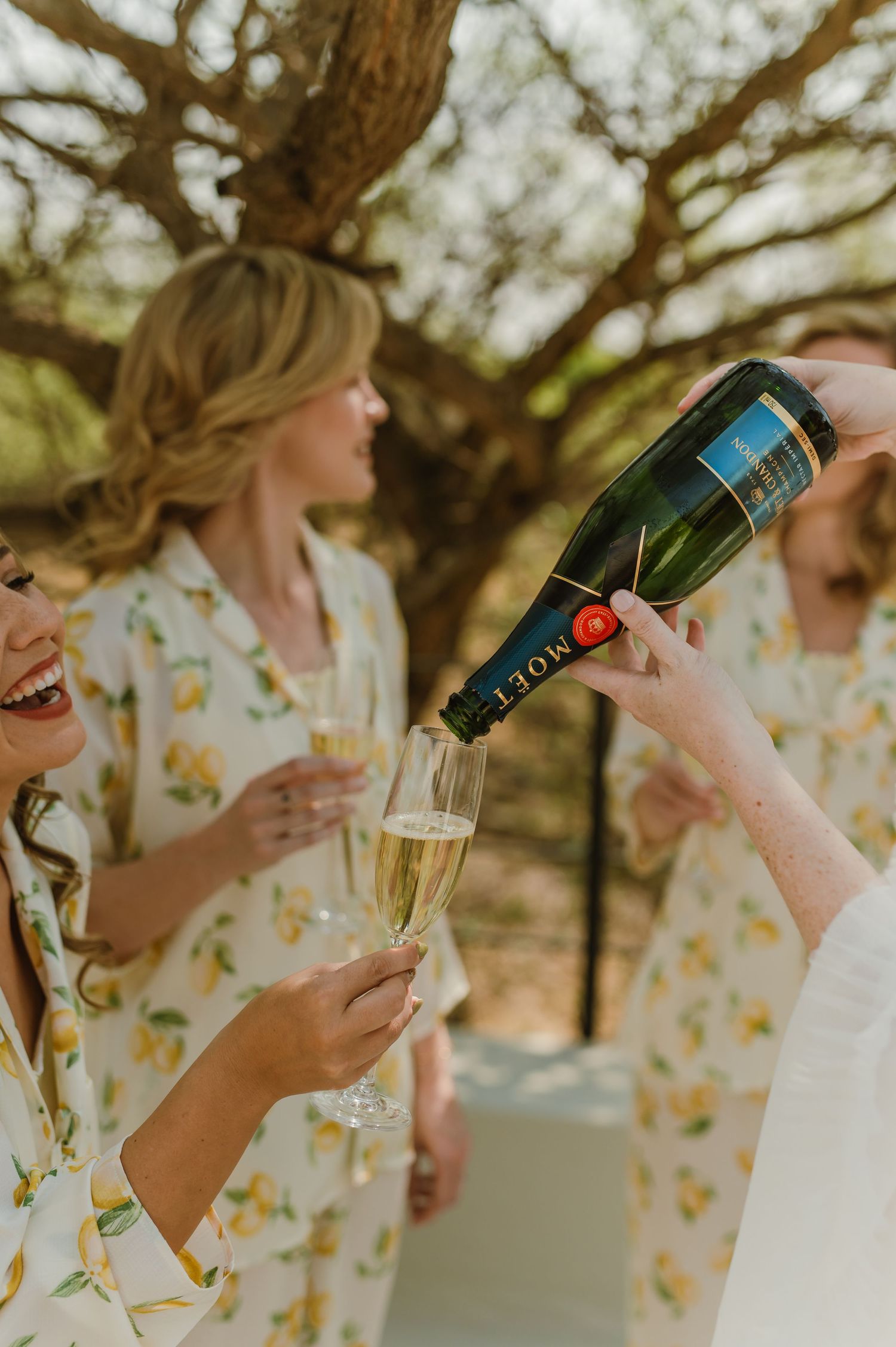 Bridesmaids in floral robes popping champagne bottles outdoors.