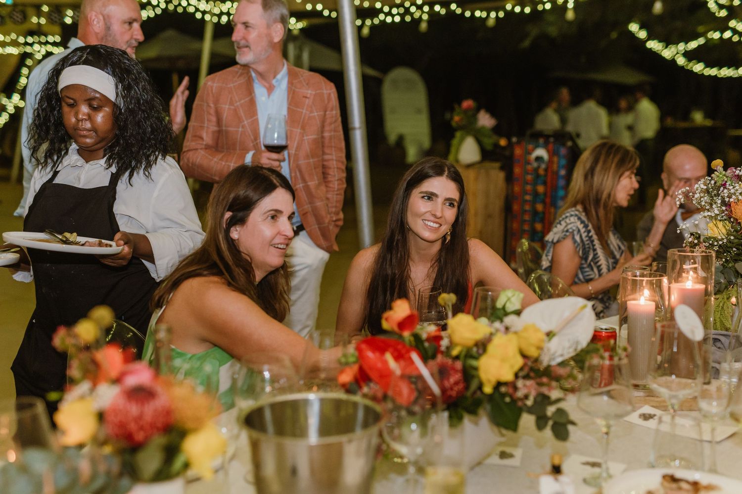Wedding guests socialize and dine at decorated tables under string lights during an evening reception.