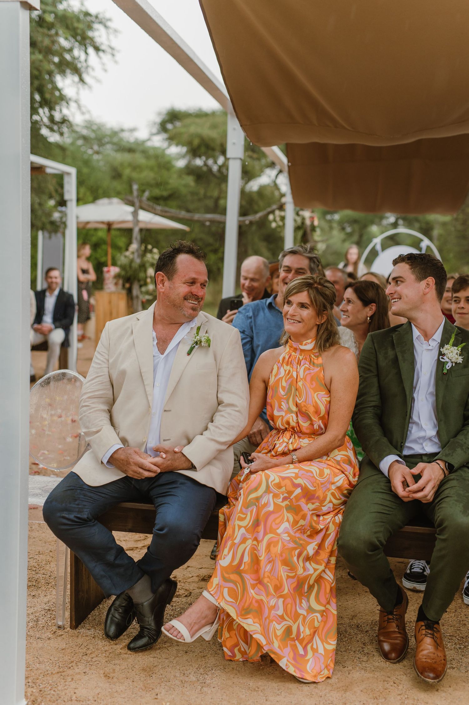 Wedding guests in colorful attire seated on benches during outdoor ceremony.