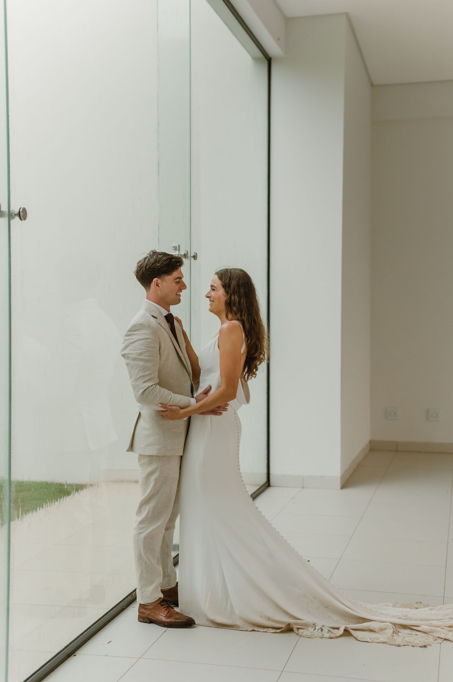 A wedding couple poses by a floor-to-ceiling window in a modern white interior.