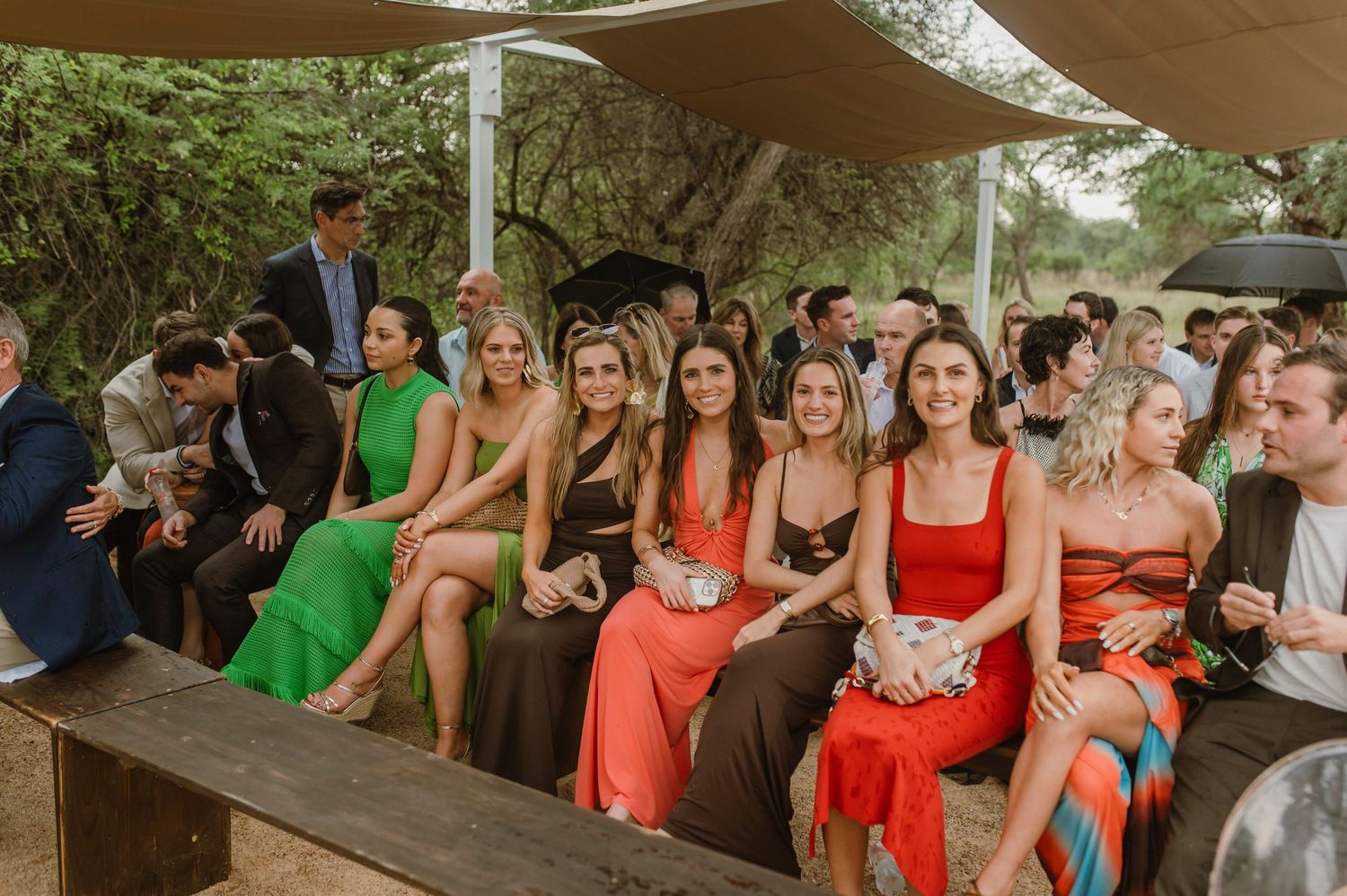 Wedding party in coral and green dresses lined up under beige shade sails at an outdoor venue.