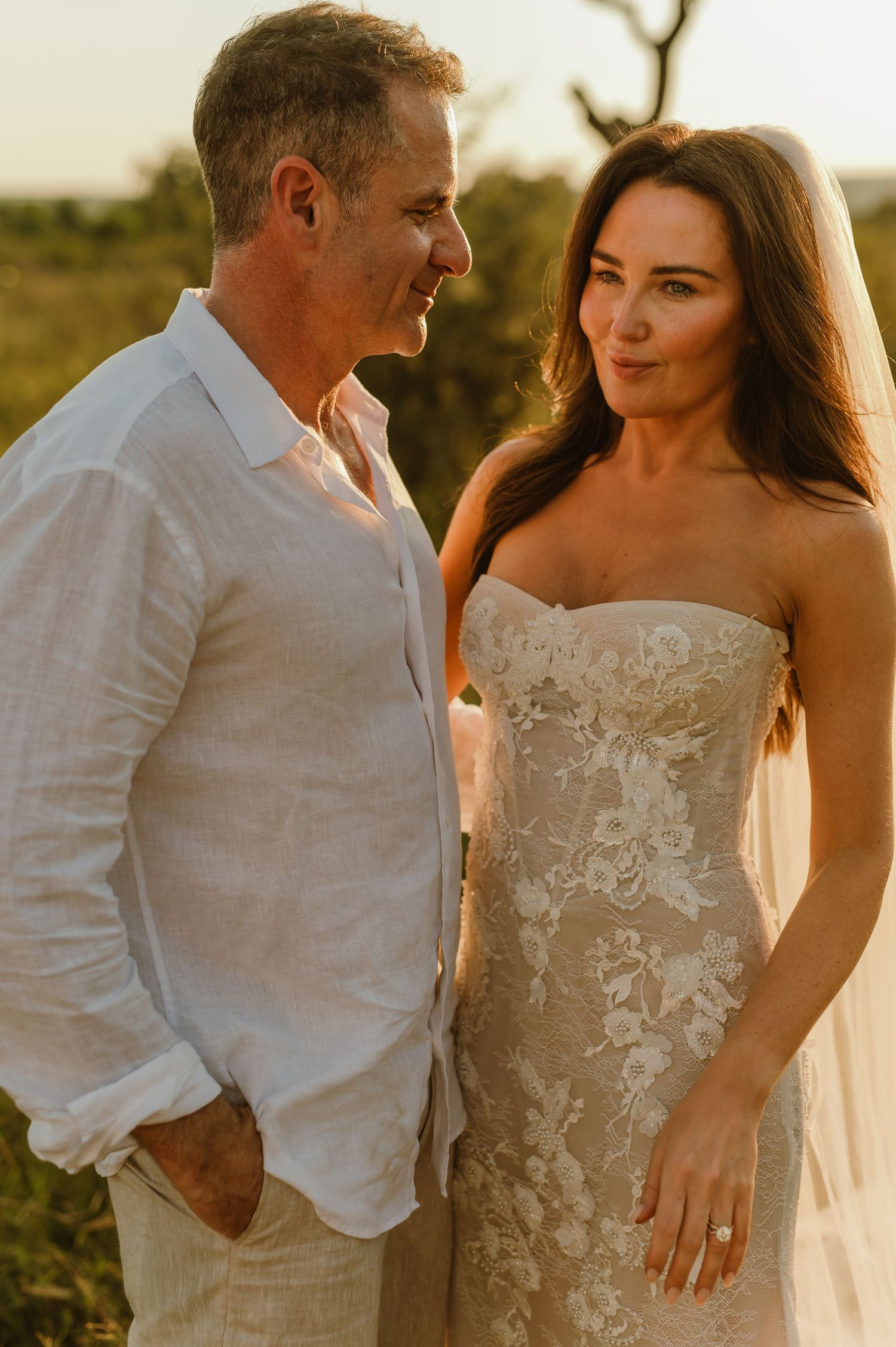 Couple in wedding attire sharing a romantic moment at golden hour, bride in lace strapless gown with veil, groom in white linen shirt.
