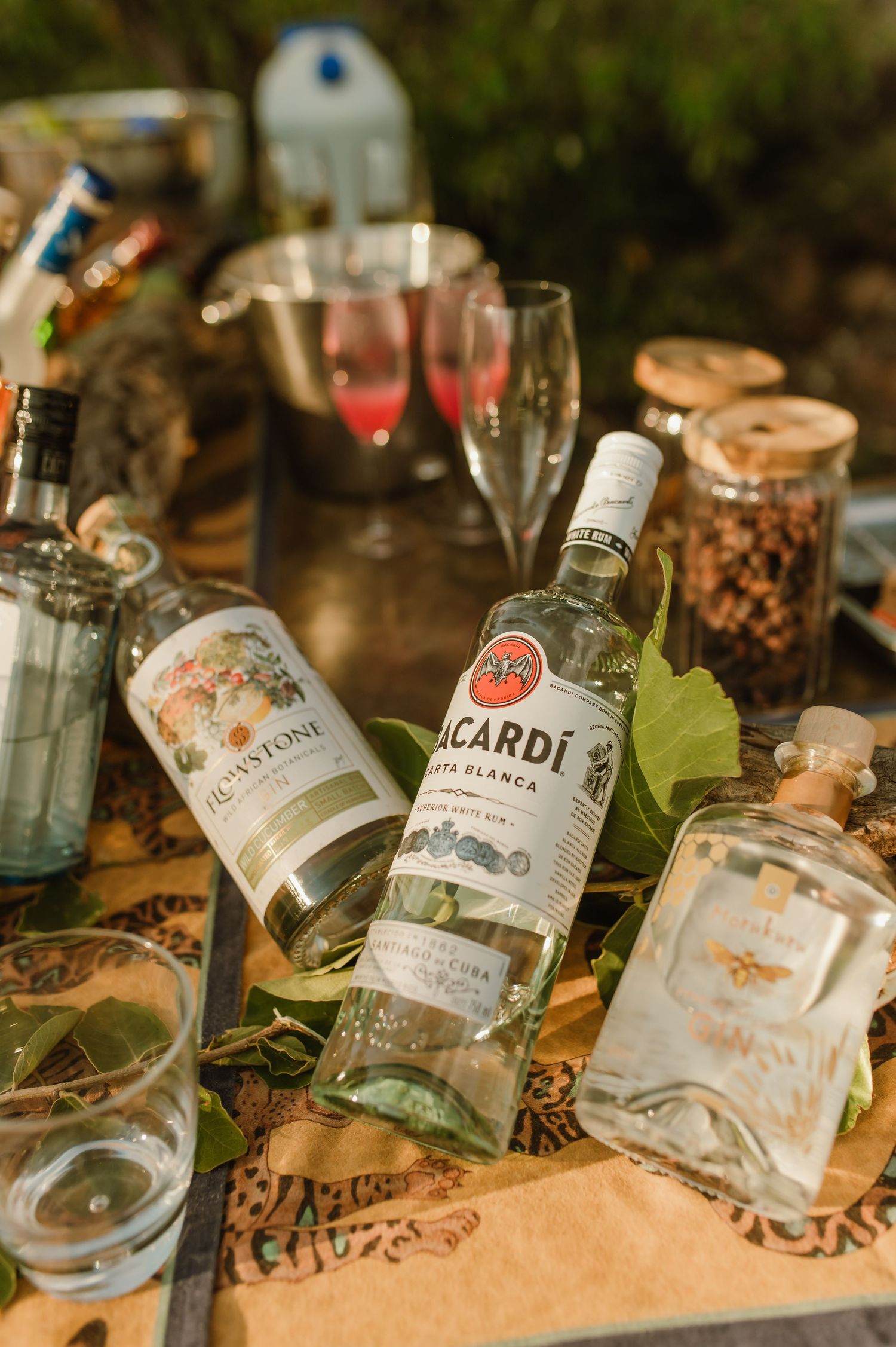 Bacardi Carta Blanca rum bottle laid on a rustic outdoor table with cocktails, glassware, and botanicals.