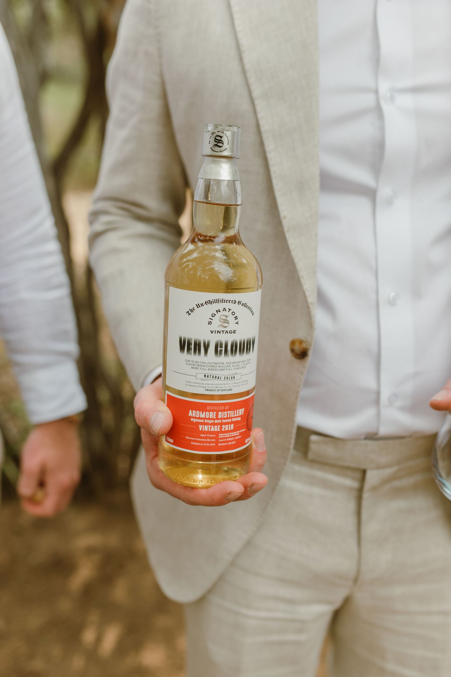 Person in light colored suit holding bottles of premium whiskey against natural background.