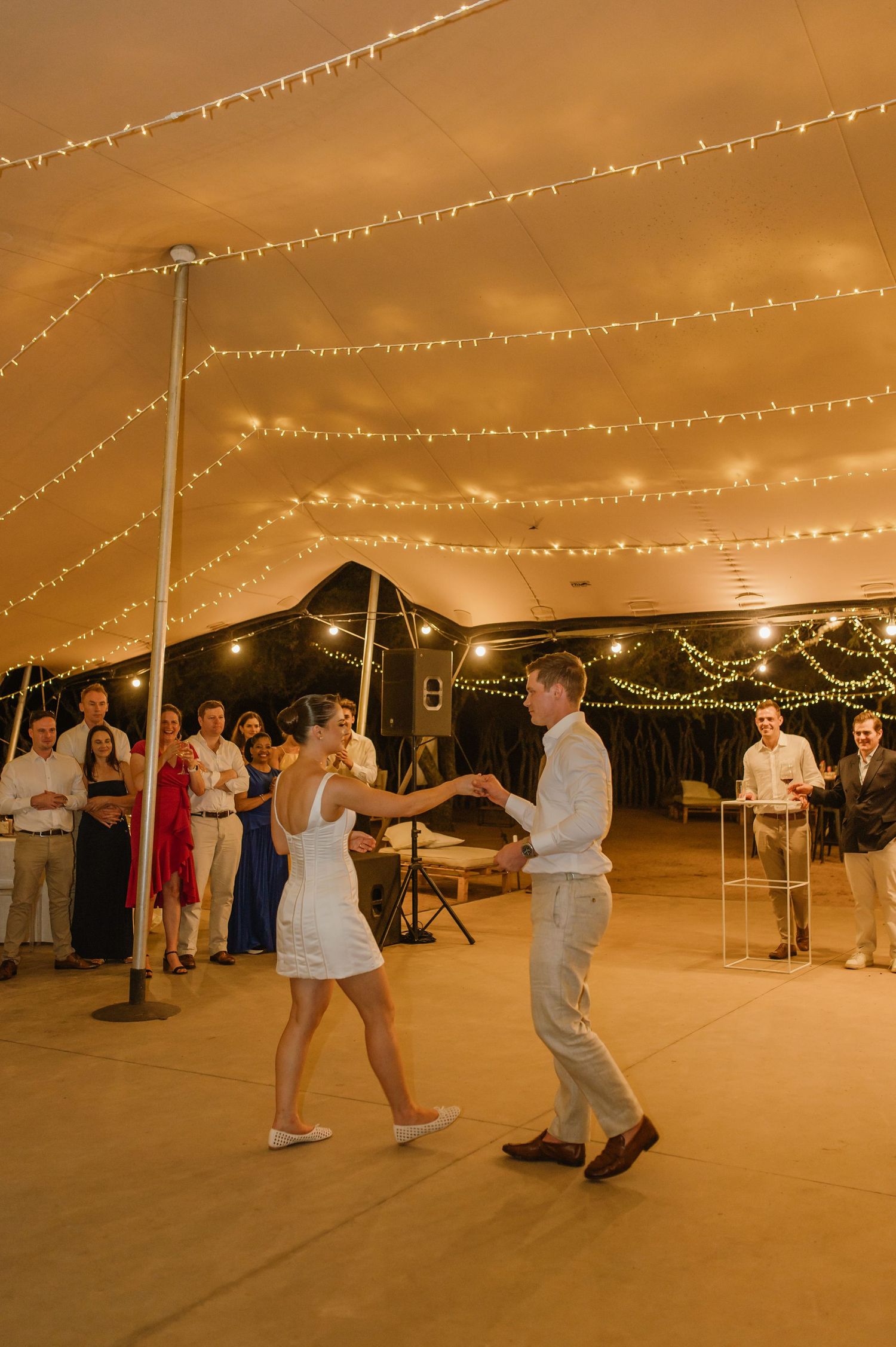 Wedding guests dancing on an outdoor dance floor under string lights at dusk.