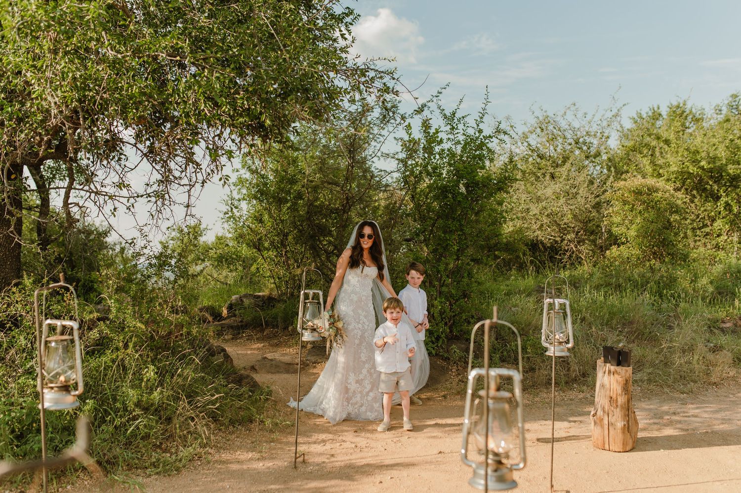 Bride in lace gown walking with two young children between lanterns on sandy outdoor aisle.