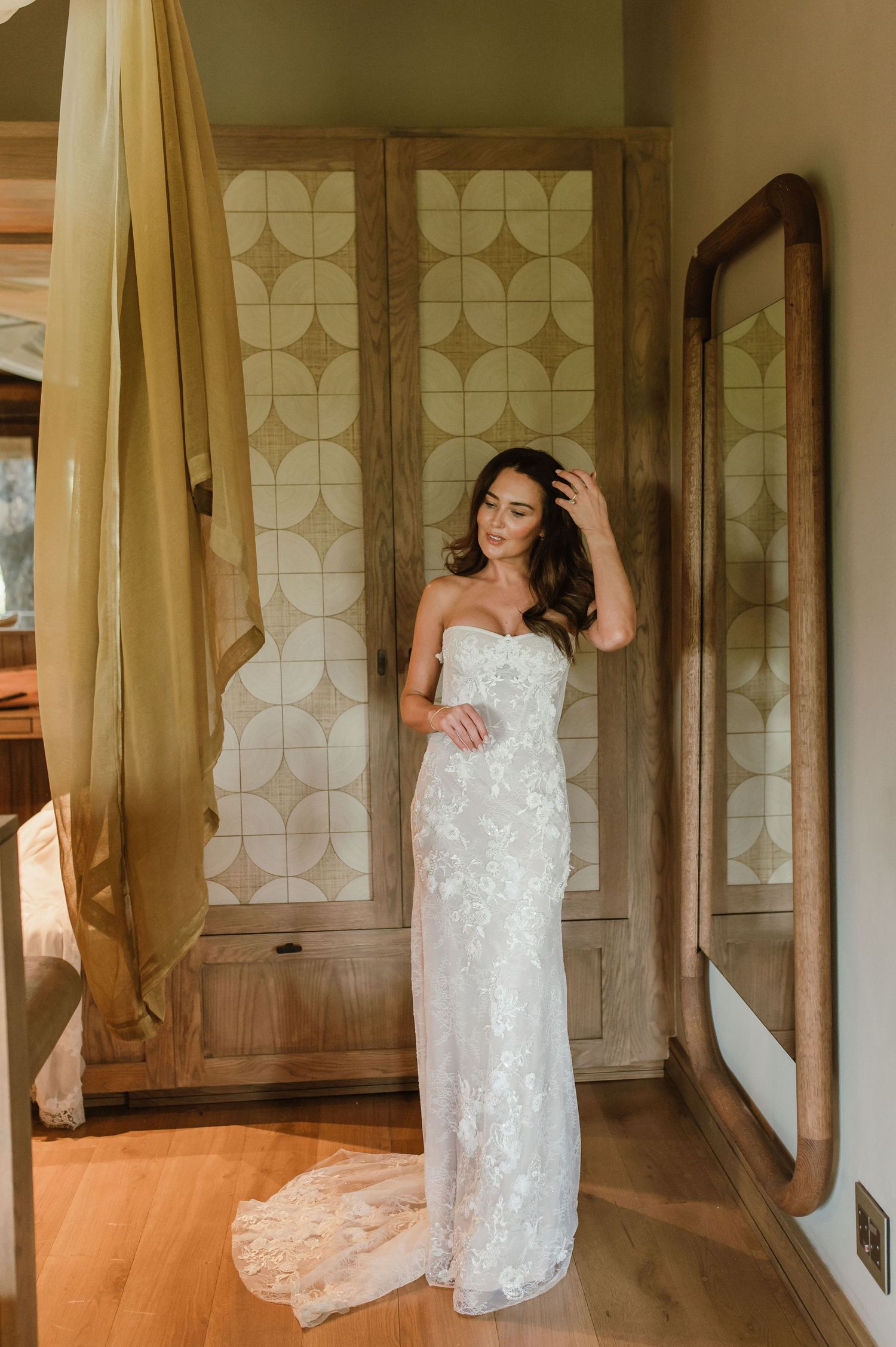 Elegant bride in strapless floral lace wedding dress posing before decorative wooden doors and mirror.