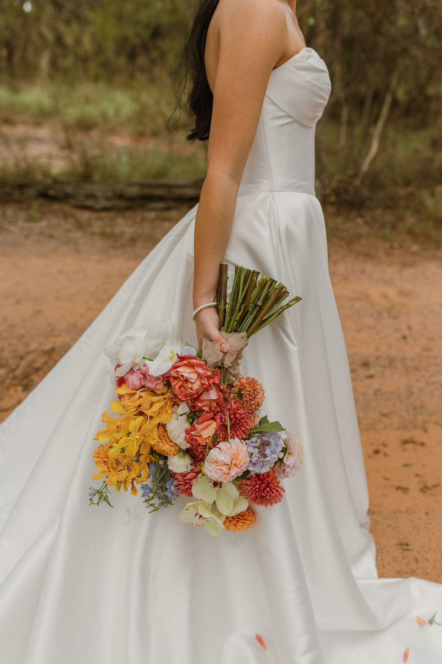 Bride holding a vibrant colorful bouquet of yellow, pink, and orange flowers in white gown.