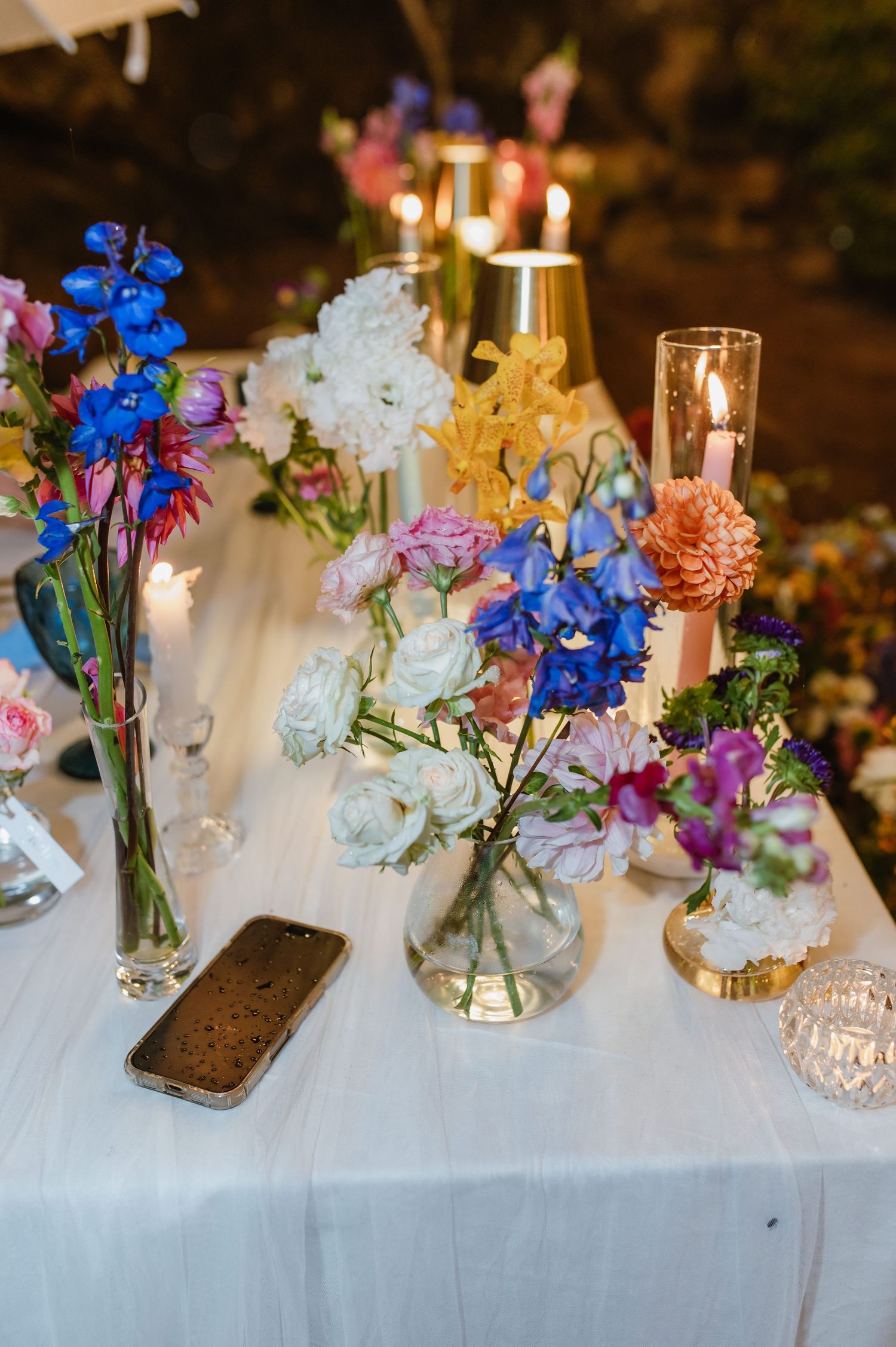 Vibrant mixed wildflower arrangements with candles and gold vases on white wedding table.