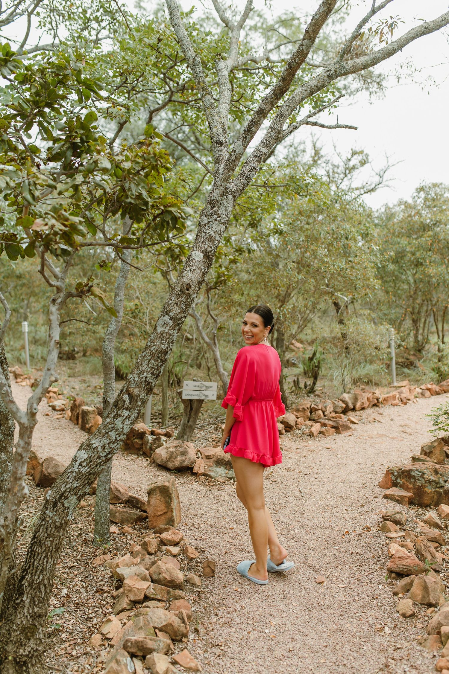 Woman in red mini dress standing on a rocky garden path surrounded by African bush trees.